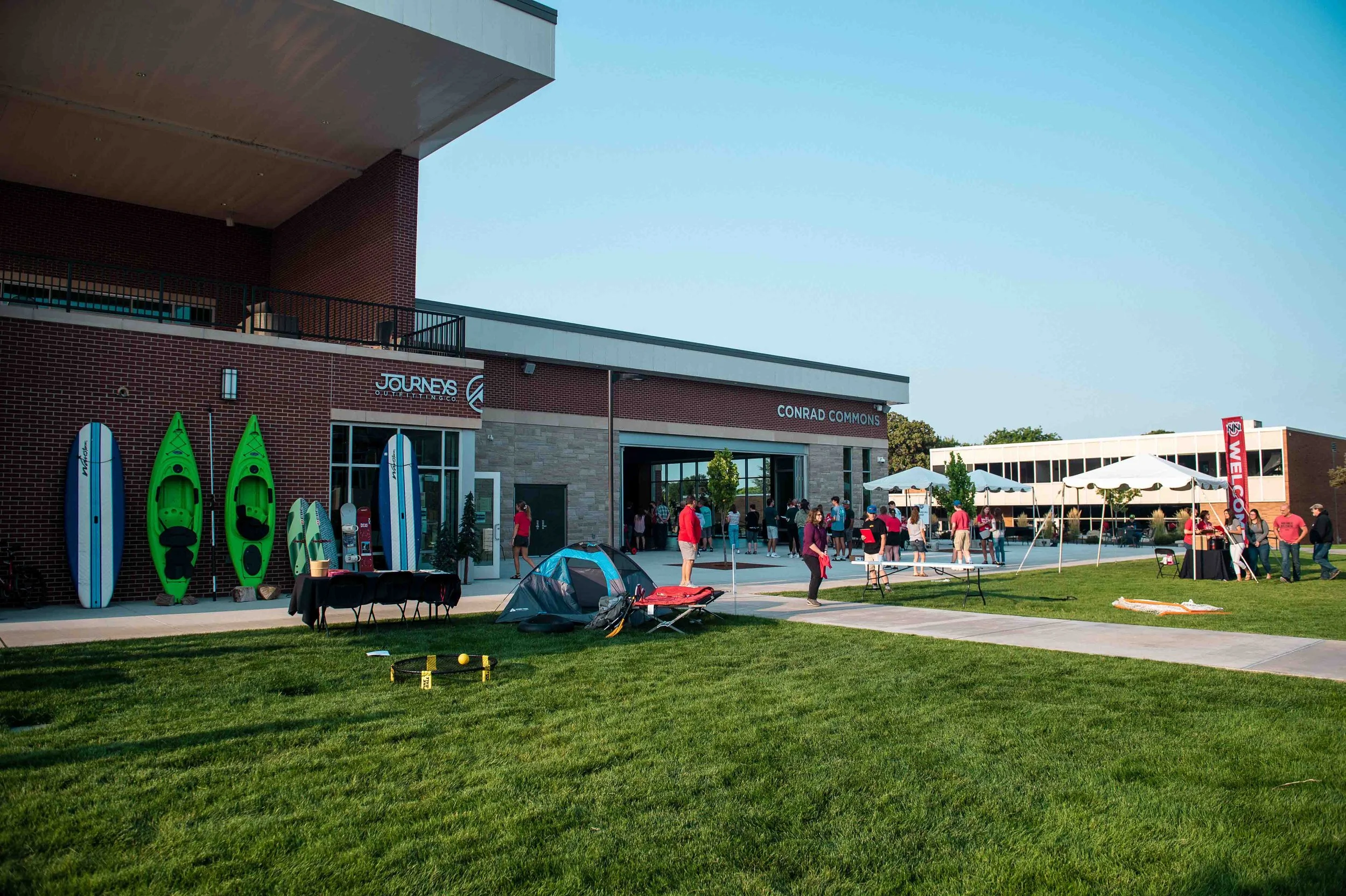 Outdoor gathering at night at a stadium with tents, kayaks, paddleboards, chairs, and people socializing.