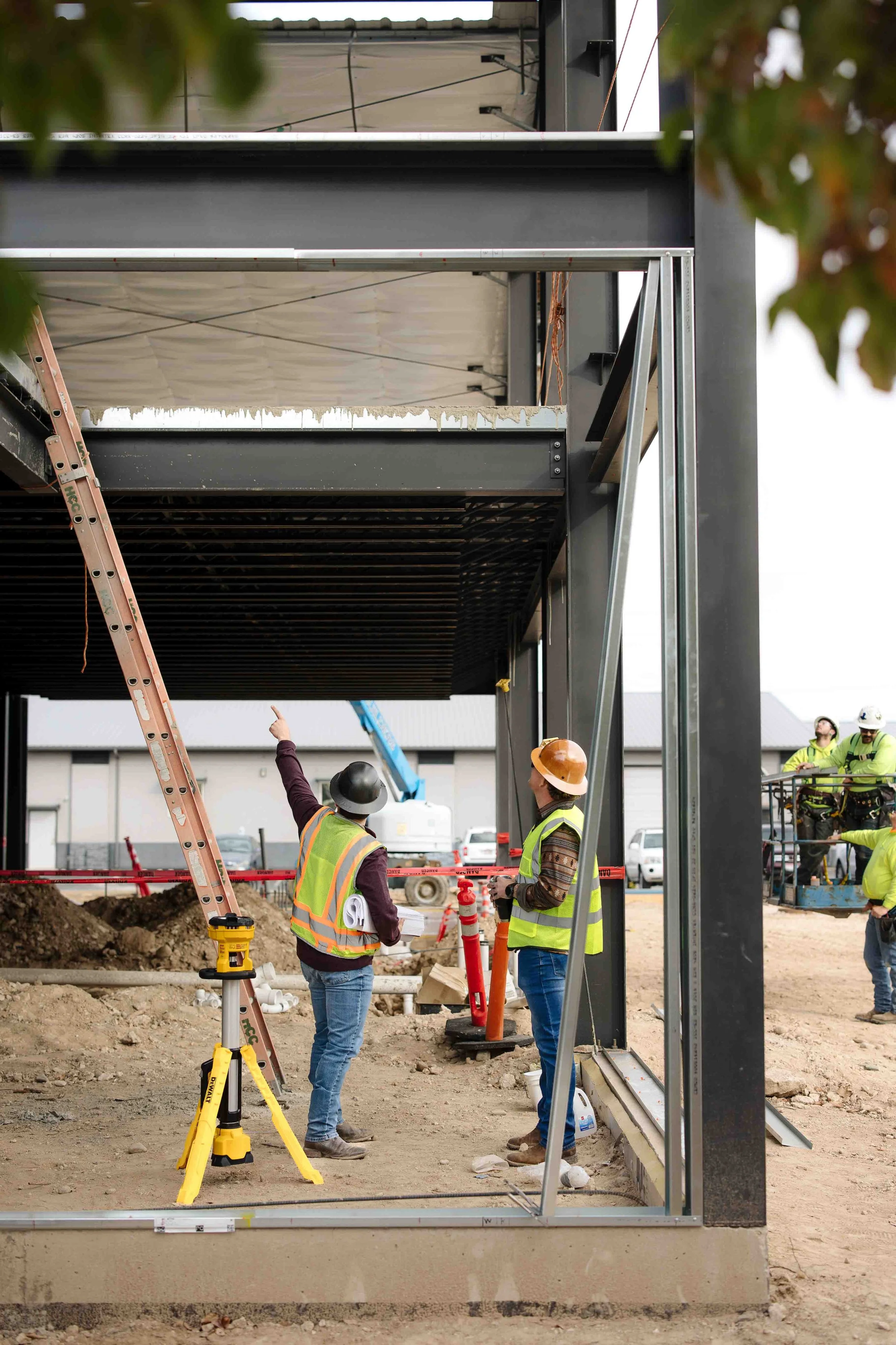 Construction workers wearing safety vests and helmets discussing at a job site with steel framework and construction equipment.