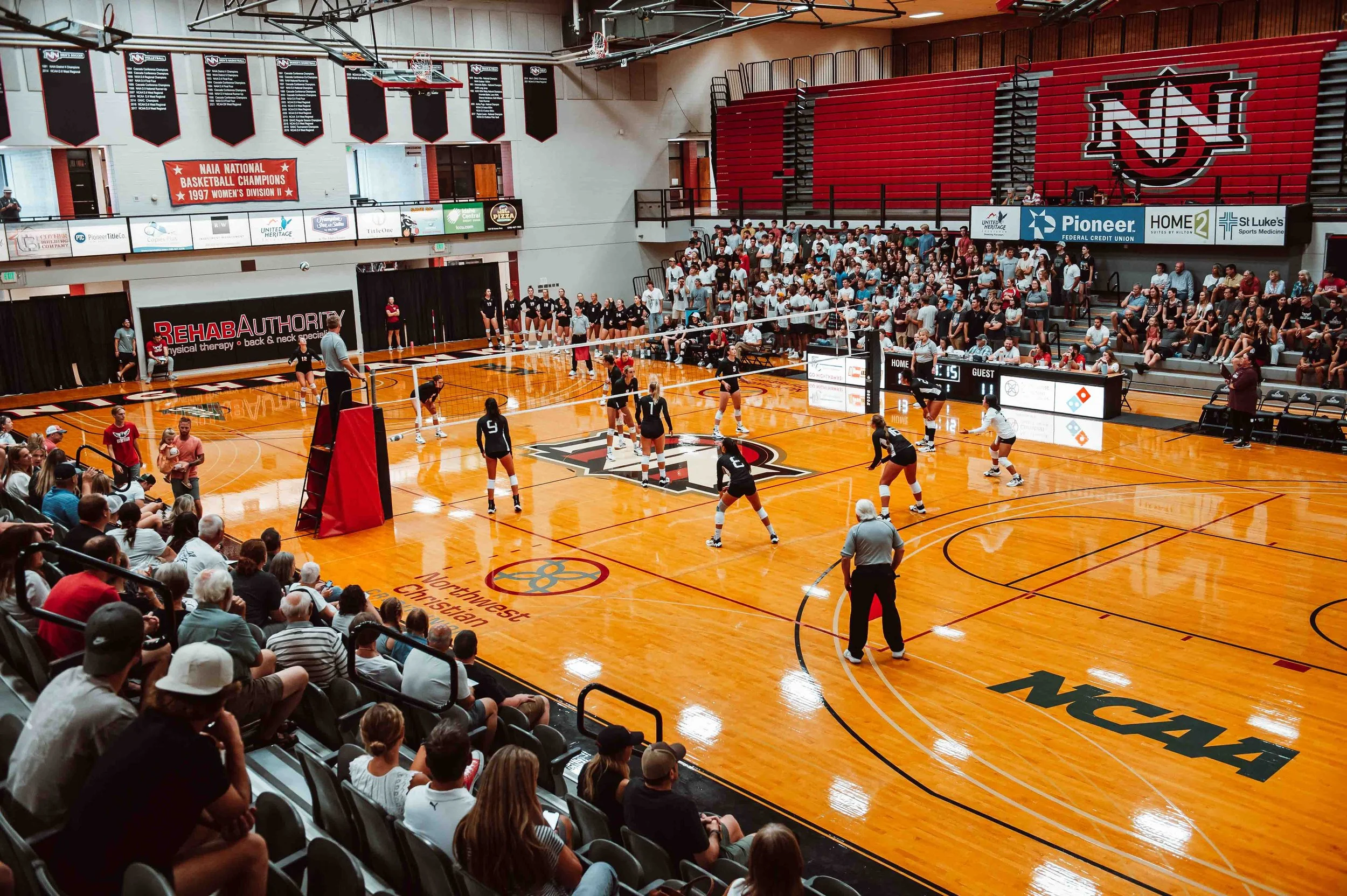An indoor volleyball game underway at a sport arena with spectators seated on both sides of the court. The court is wooden with the NCAA logo, and players are positioned for service or defense, with referees and scorekeepers present.