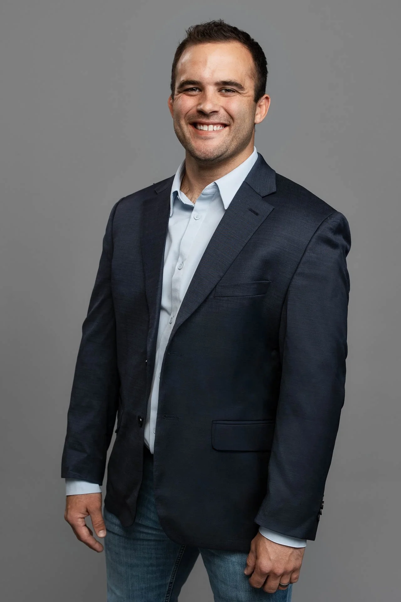 A man smiling, wearing a dark suit jacket and jeans, standing against a gray background.