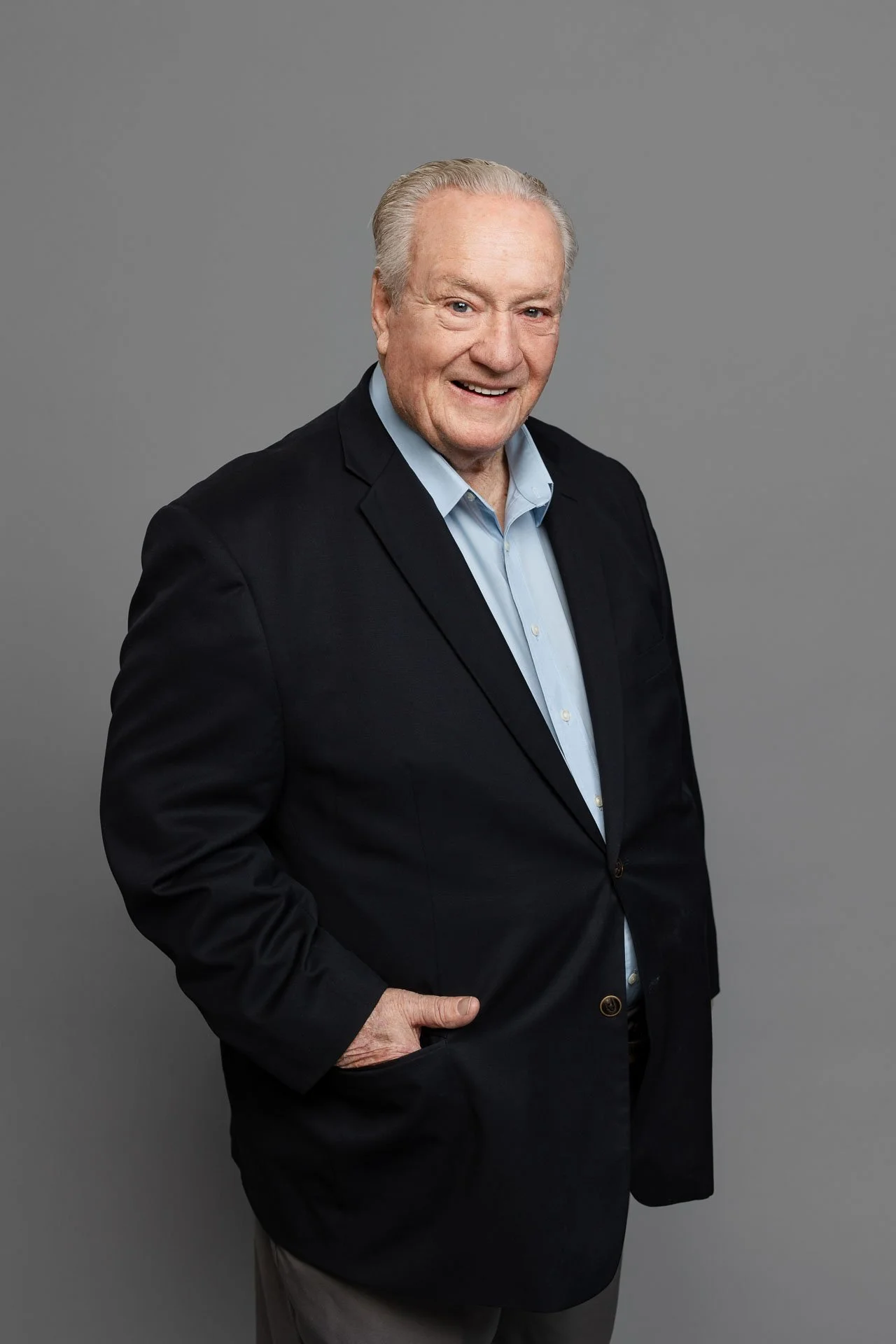 A senior man in a black blazer and light blue shirt smiling at the camera, standing against a plain gray background.