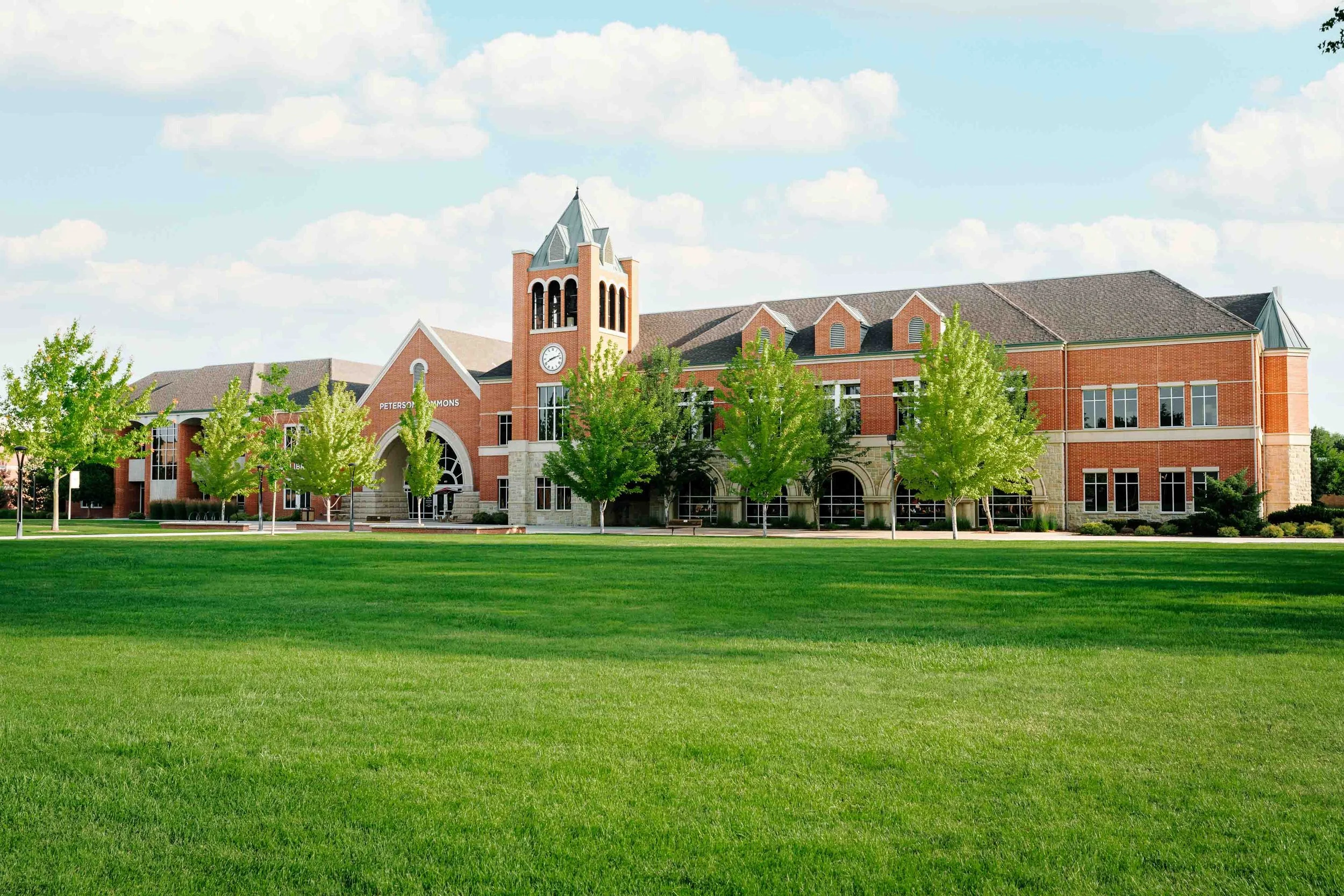 A large brick school building with a clock tower, trees in front, and a well-maintained lawn in the foreground under a partly cloudy sky.