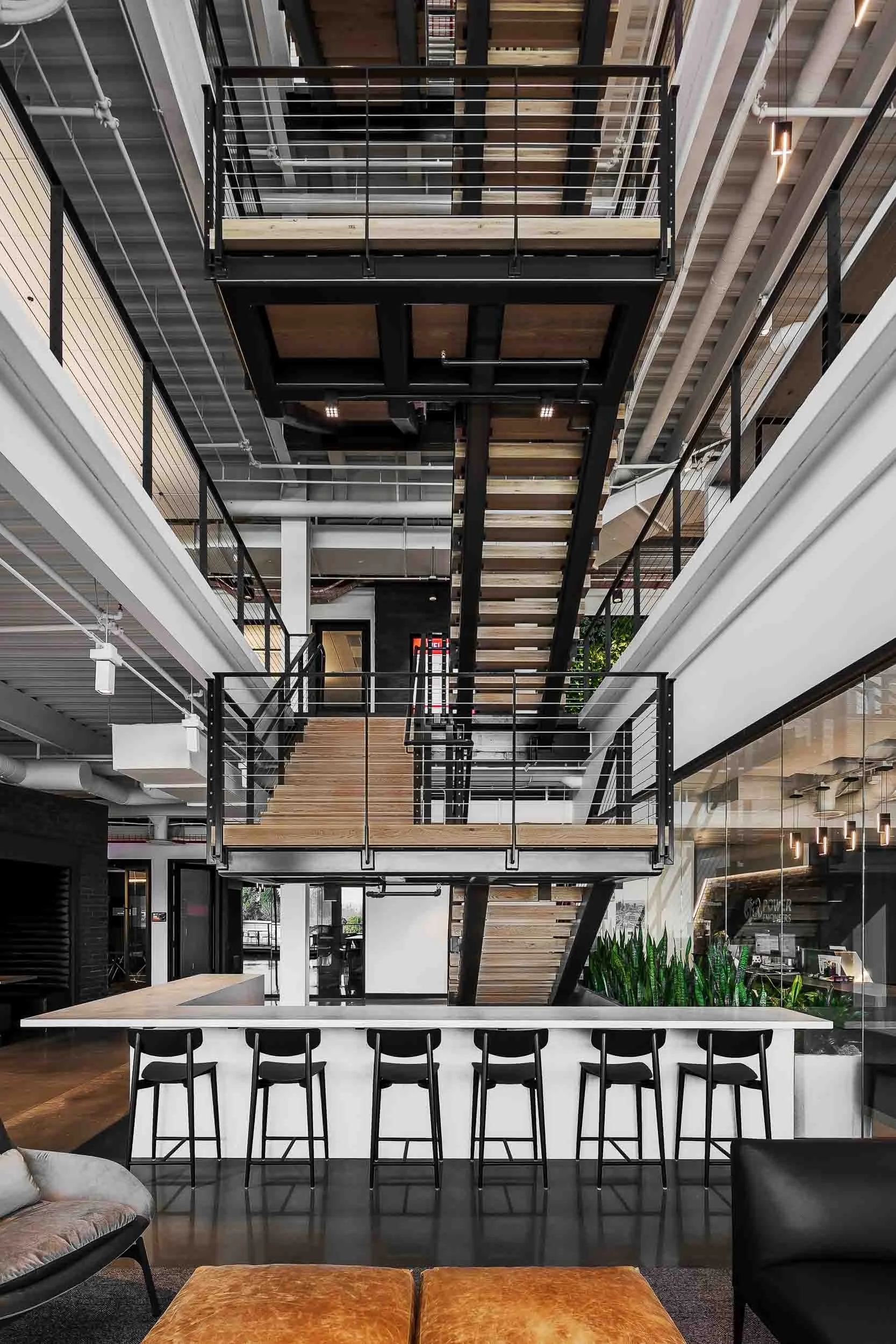 A modern multi-level office interior featuring a staircase with wooden steps and black metal supports, surrounded by glass walls and railings, with seating and a bar counter with five black chairs in the foreground.