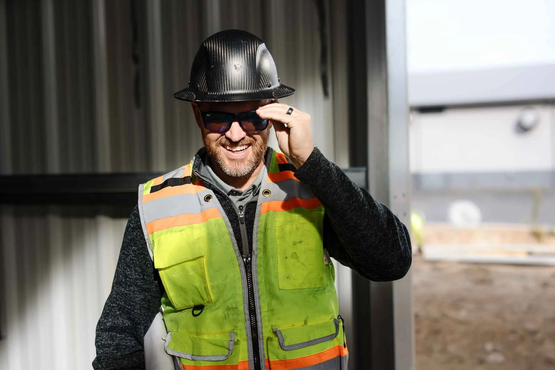 A construction worker wearing a black hard hat, sunglasses, a gray hoodie, and a neon yellow safety vest smiling and adjusting his sunglasses at a building site.