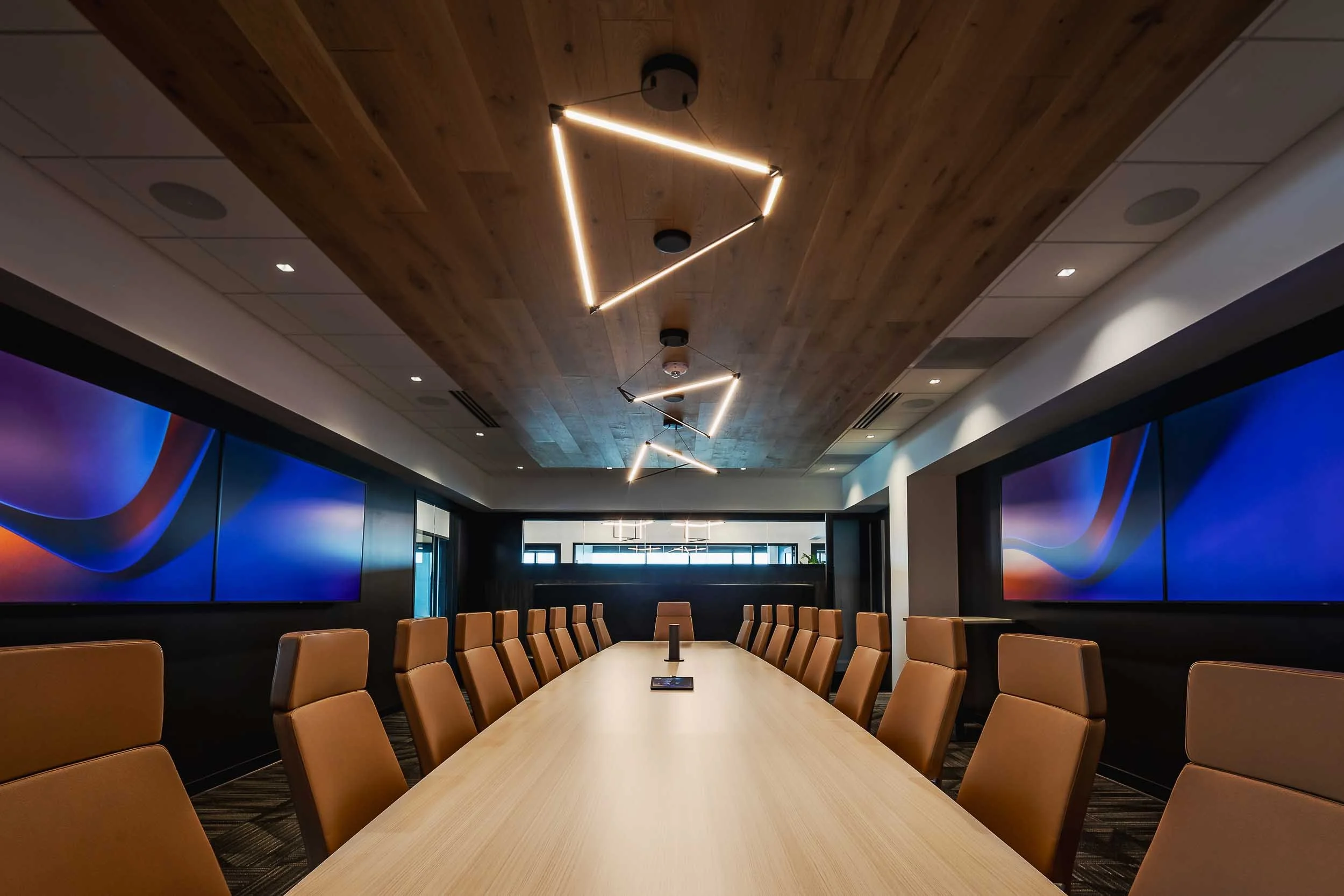 A modern conference room with a long wooden table, brown leather chairs, large video screens on the walls, and unique geometric LED lighting fixtures on the wooden ceiling.