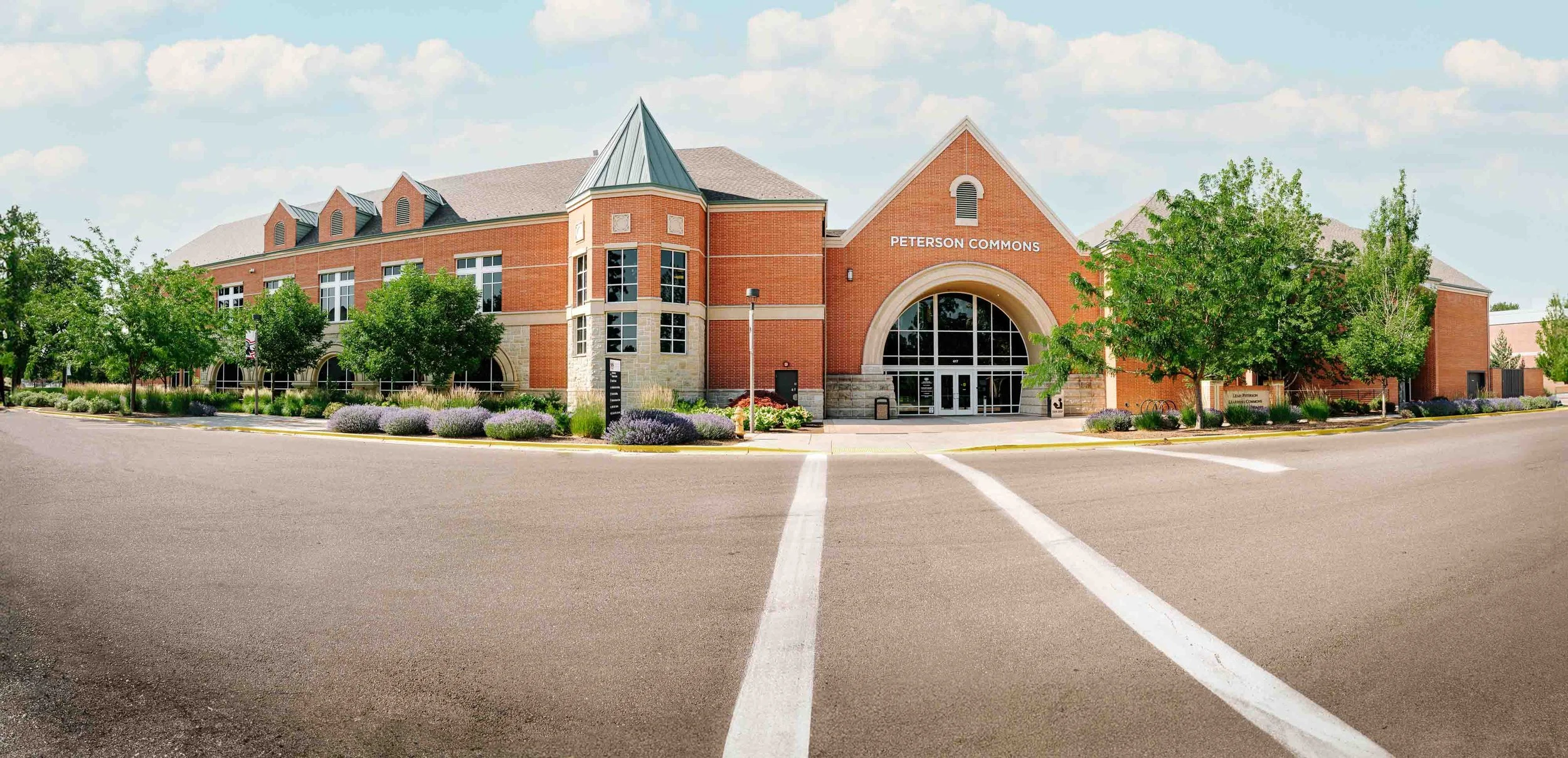 Front view of Peterson Commons, a large brick building with a prominent arched entrance, surrounded by trees and landscaped bushes, with a clear sky overhead.