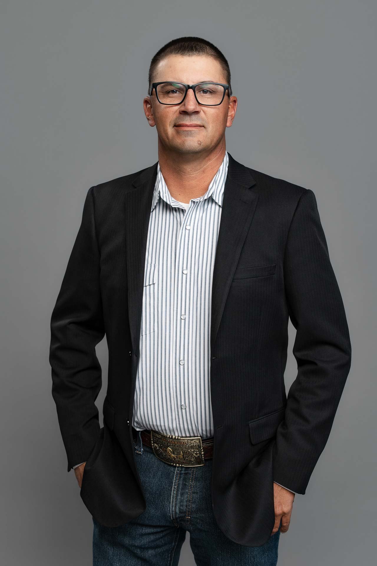 A man wearing glasses, a black blazer, a striped button-up shirt, and jeans, standing against a gray background.