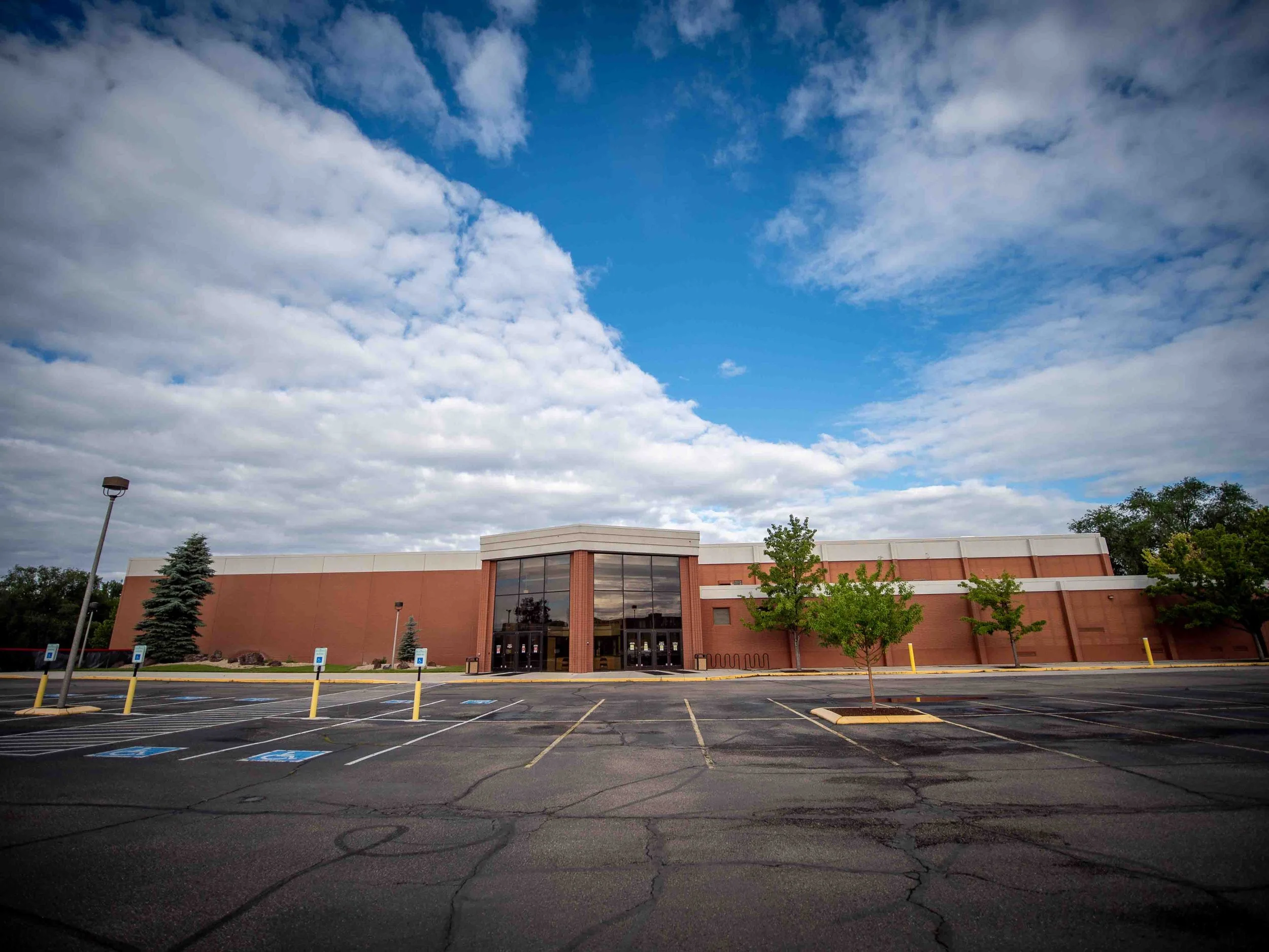 Empty parking lot in front of a large retail store with a red brick exterior, glass entrance, and small trees, under a partly cloudy sky.