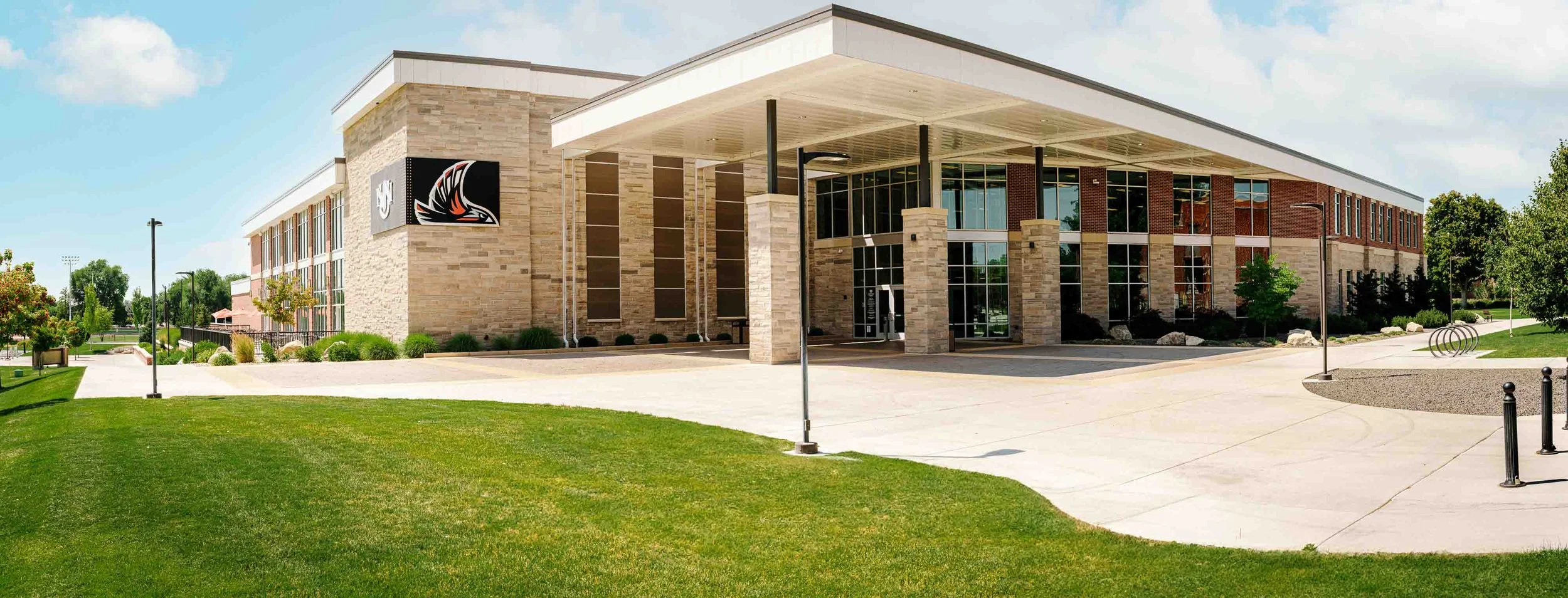 Modern school building with brick and stone exterior, large glass windows, and a covered entrance, surrounded by a well-maintained lawn and trees under a blue sky.