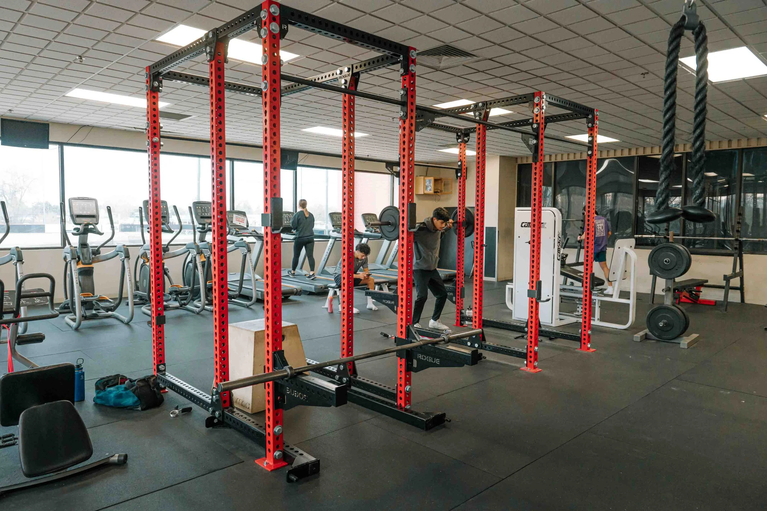 People working out with gym equipment including treadmills, a squat rack, and weightlifting machines in a fitness center.