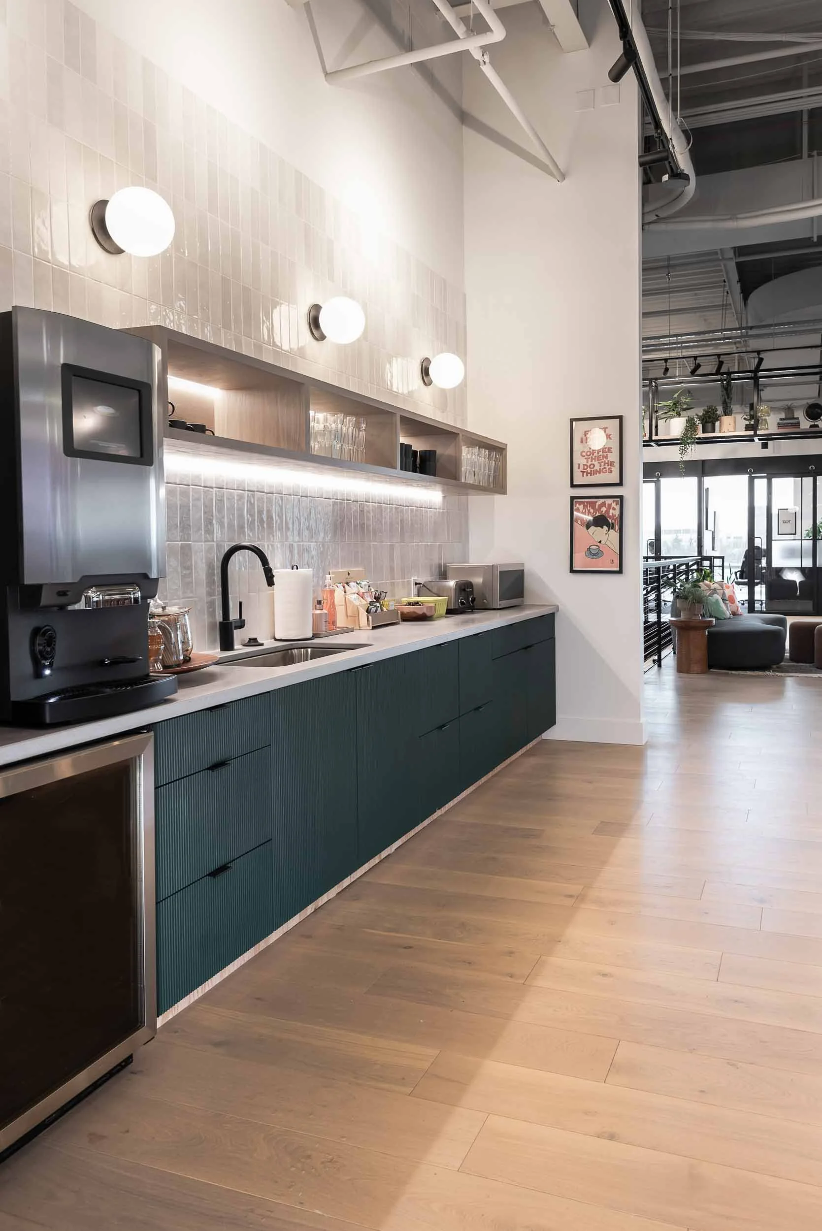 A modern kitchen area with a dark green lower cabinet, a white countertop, and wall-mounted lighting fixtures. There are various small appliances and items on the counter, and an open shelf with glassware and other items. The space extends into a bright room with seating and large windows.