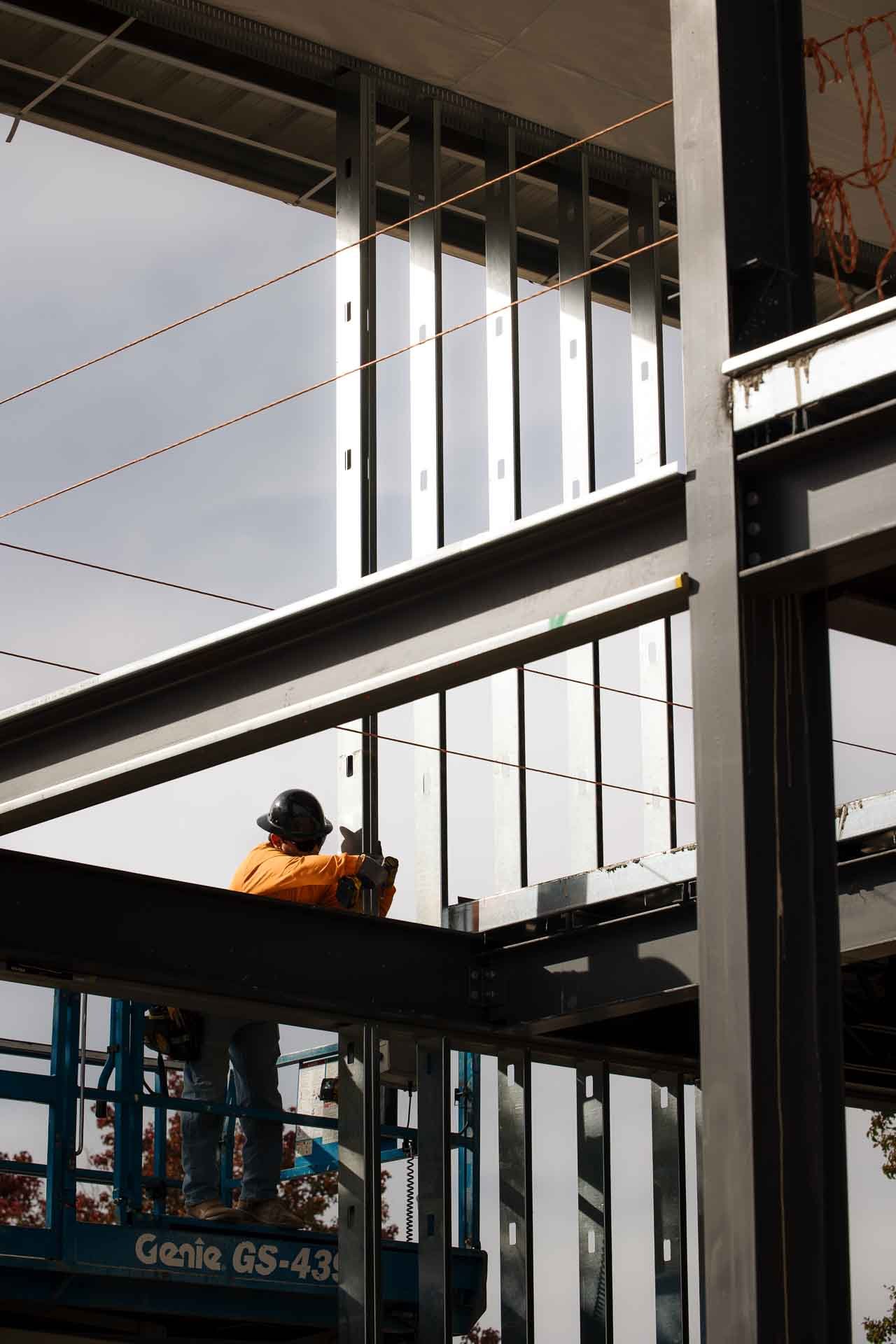 A construction worker wearing an orange jacket, safety helmet, and gloves working on the steel framework of a building during daytime.
