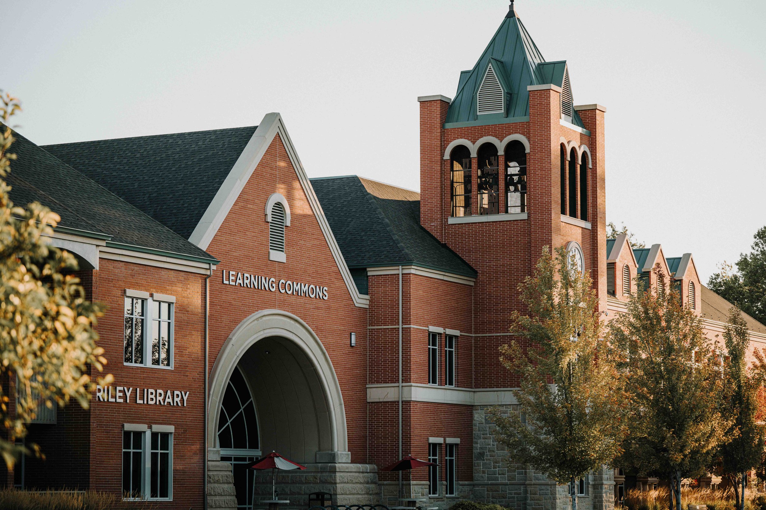 A large brick building with a steeple, labeled 'Learning Commons' and 'Riley Library', featuring arched windows and an entrance with steps and a red umbrella, surrounded by trees.