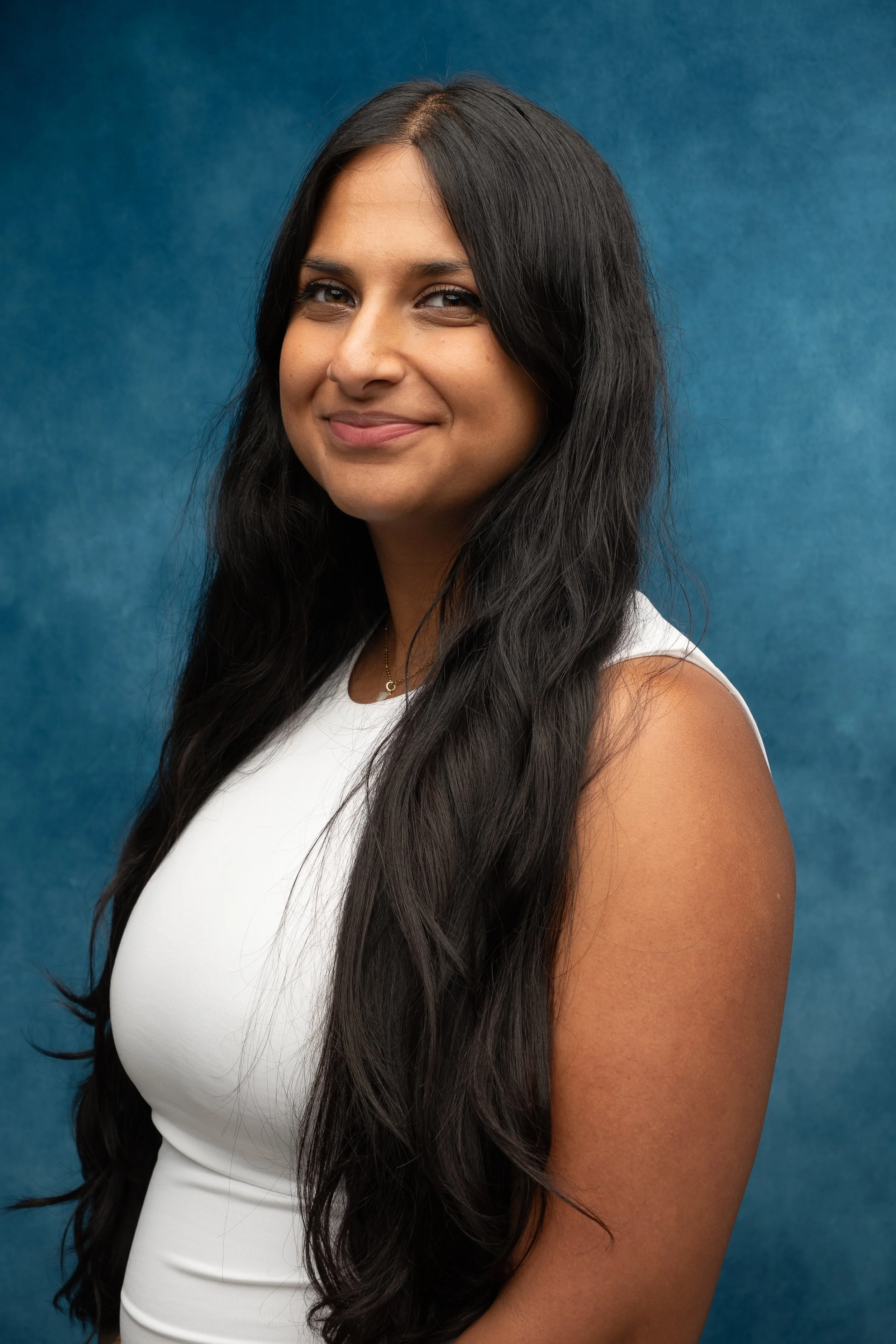A young woman with long black hair wearing a white sleeveless top, smiling slightly, against a blue background.