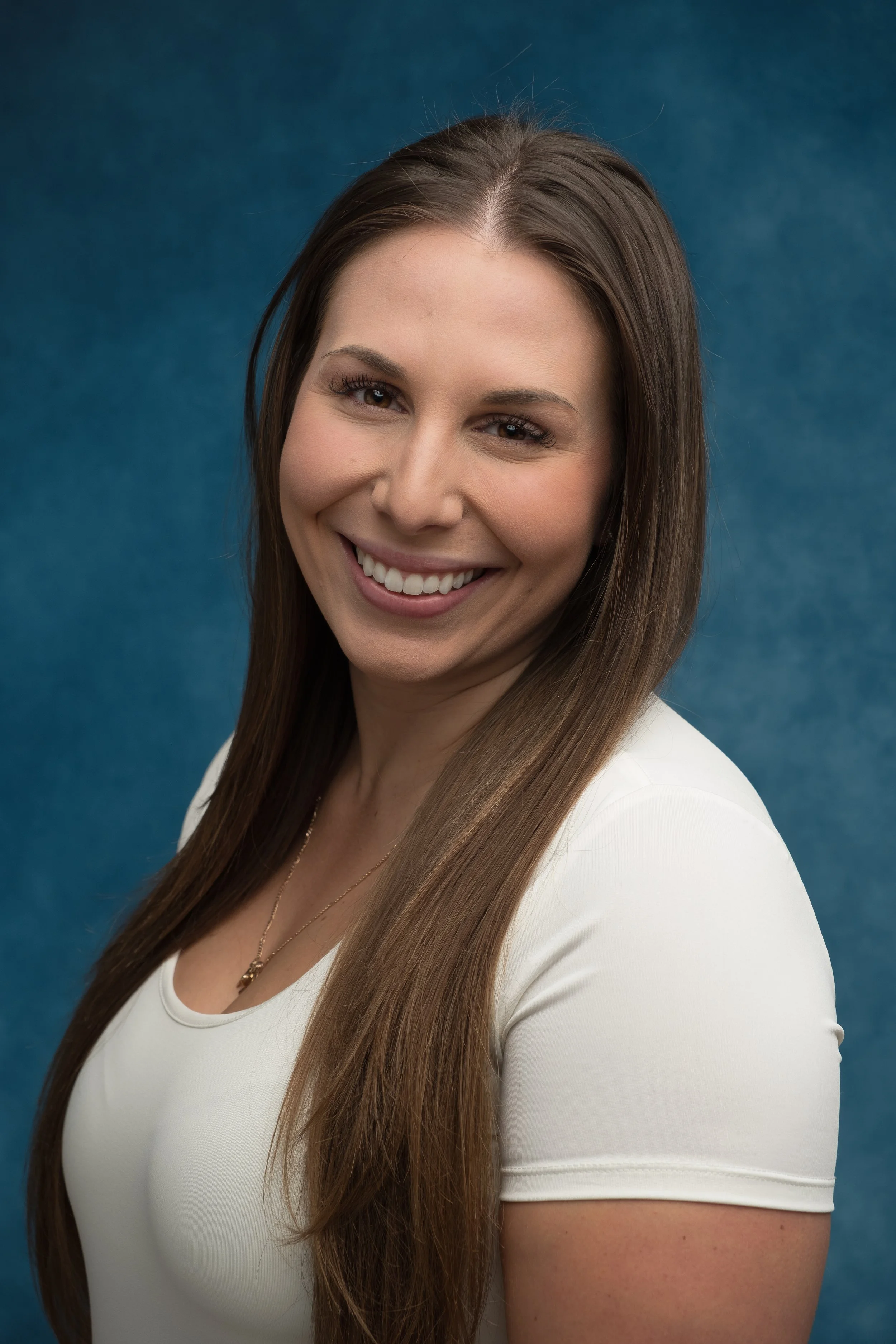 Portrait of a smiling woman with long Brown hair wearing a white top posing against a blue background.