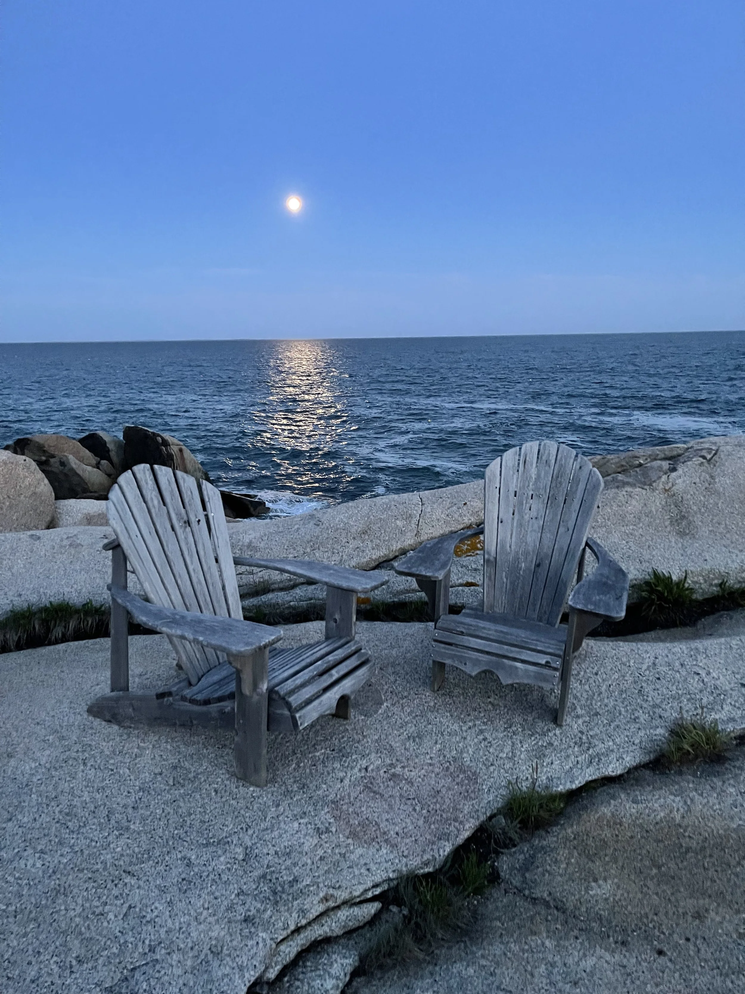 Two weathered gray wooden Adirondack chairs on a rocky beach facing the ocean at dusk, with the moon shining over the water.