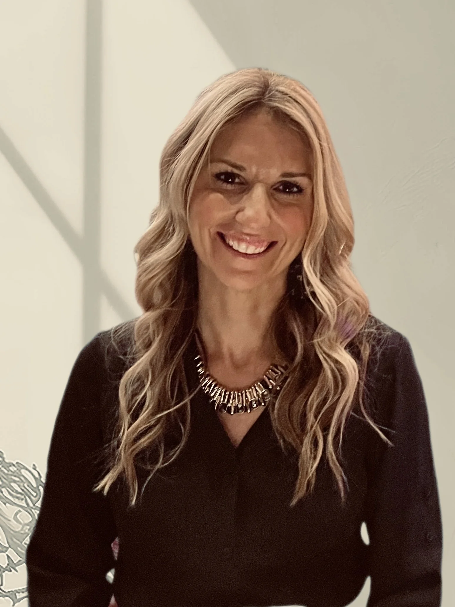 Smiling woman with blonde wavy hair, wearing a black top and a jeweled necklace, in front of a light-colored background.