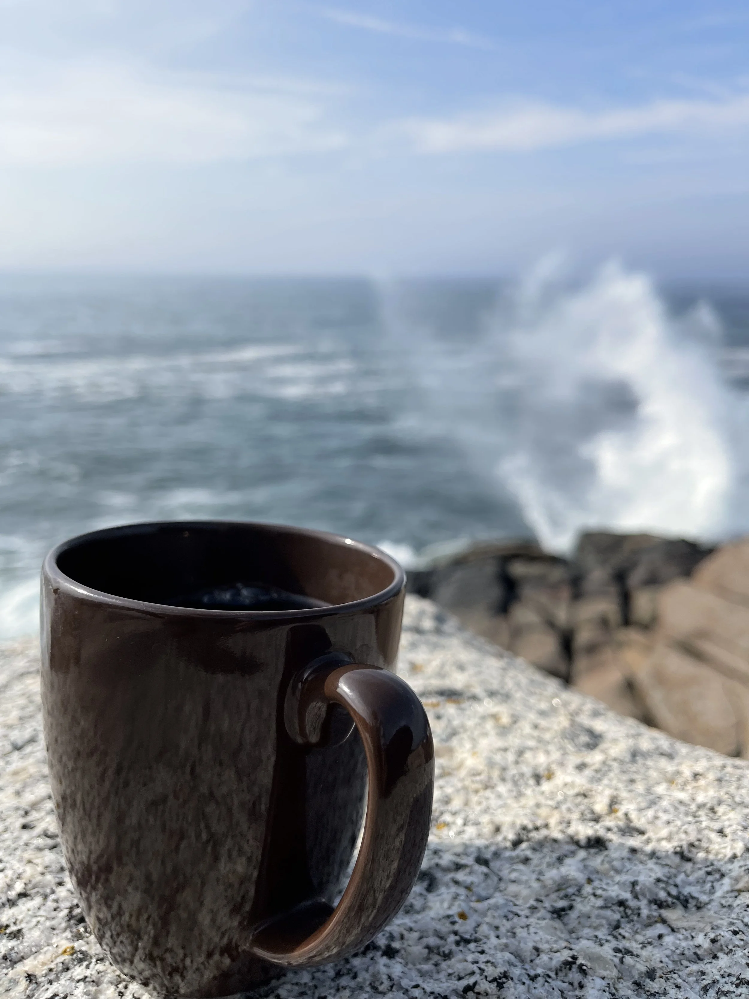 Brown ceramic coffee mug on a rocky surface with an ocean and blue sky with clouds in the background, and waves crashing against rocks.
