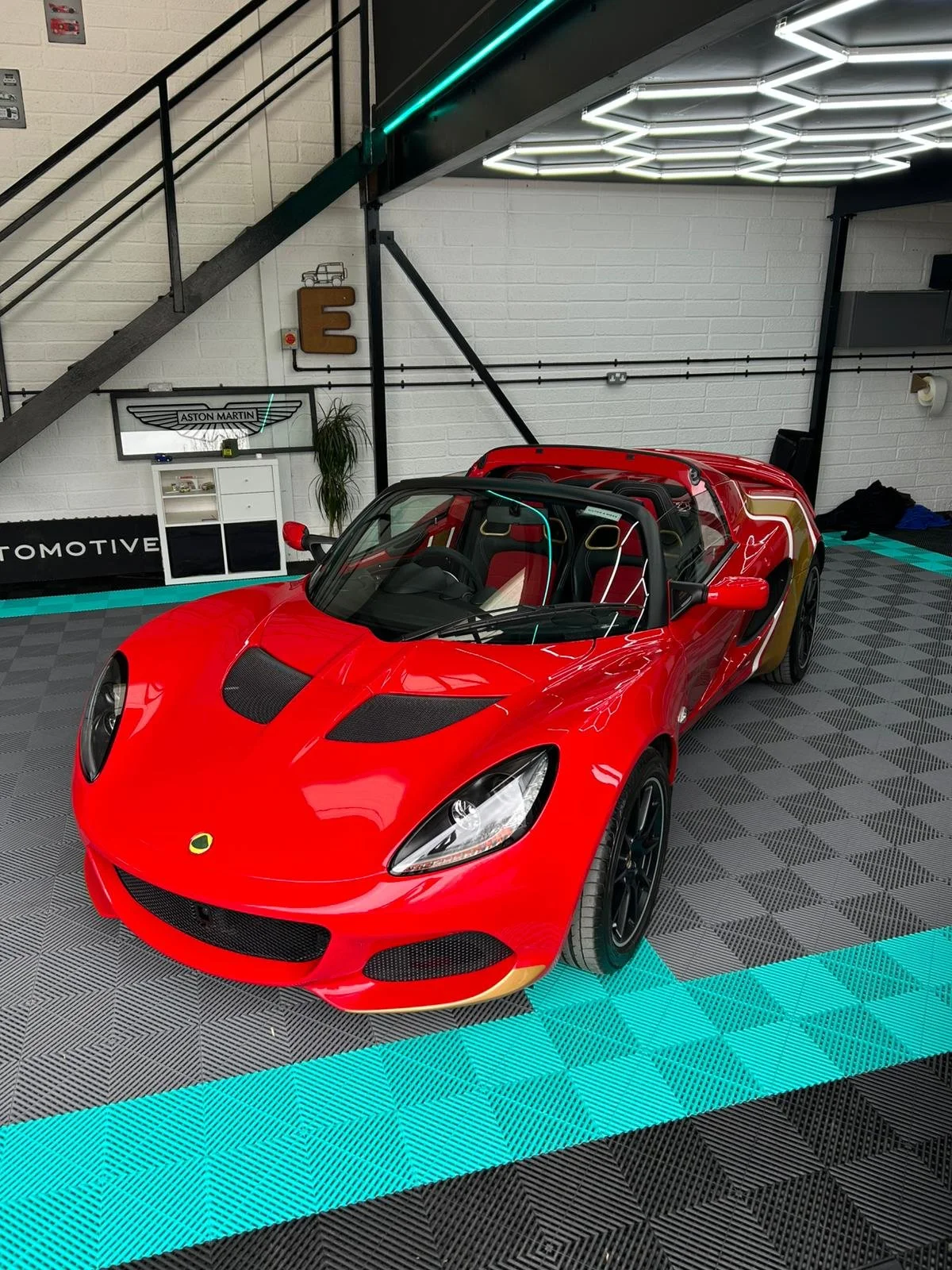 Red Lotus sports car inside a garage with white brick walls, illuminated hexagonal light ceiling, and racing seats visible through the open cockpit.