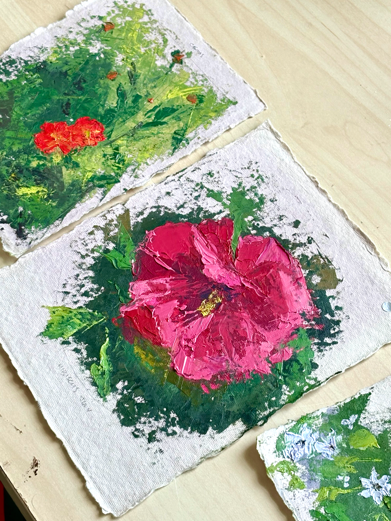 Two small canvas paintings of flowers, one with red and yellow flowers on green foliage, the other with large pink flowers and green leaves, on a light wooden surface.