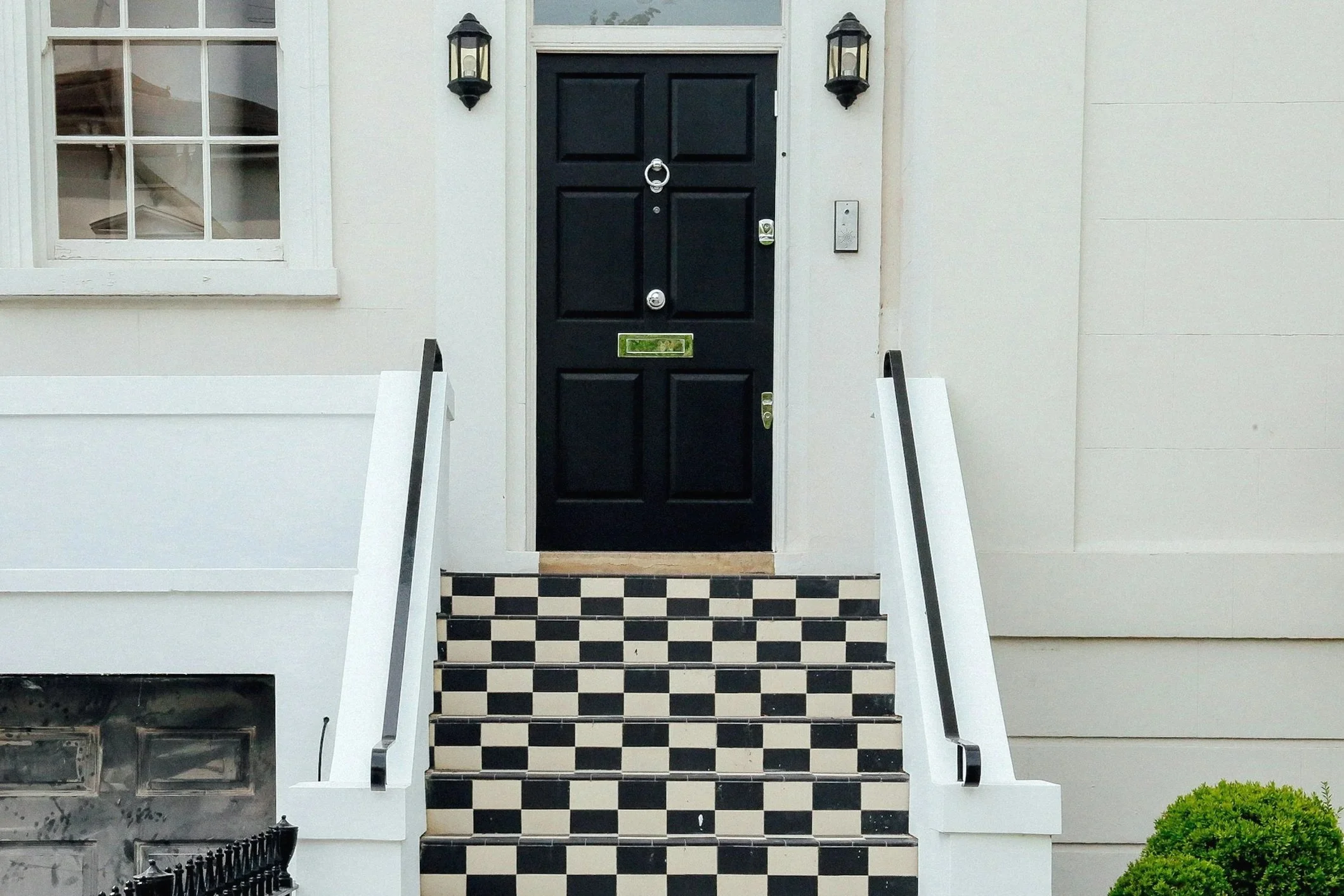 Front entrance to a house with black front door, two wall-mounted lantern style lights on either side, checkered black and white stairs, white railing, and a small green shrub at the bottom right.