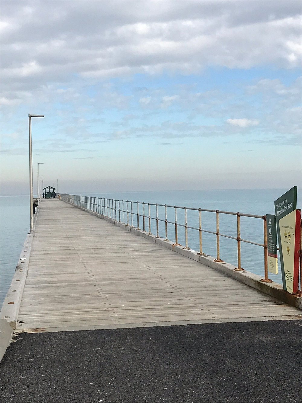 Wooden pier extending over the water with a railing on the sides and a small shelter at the end. Overcast sky with some clouds.