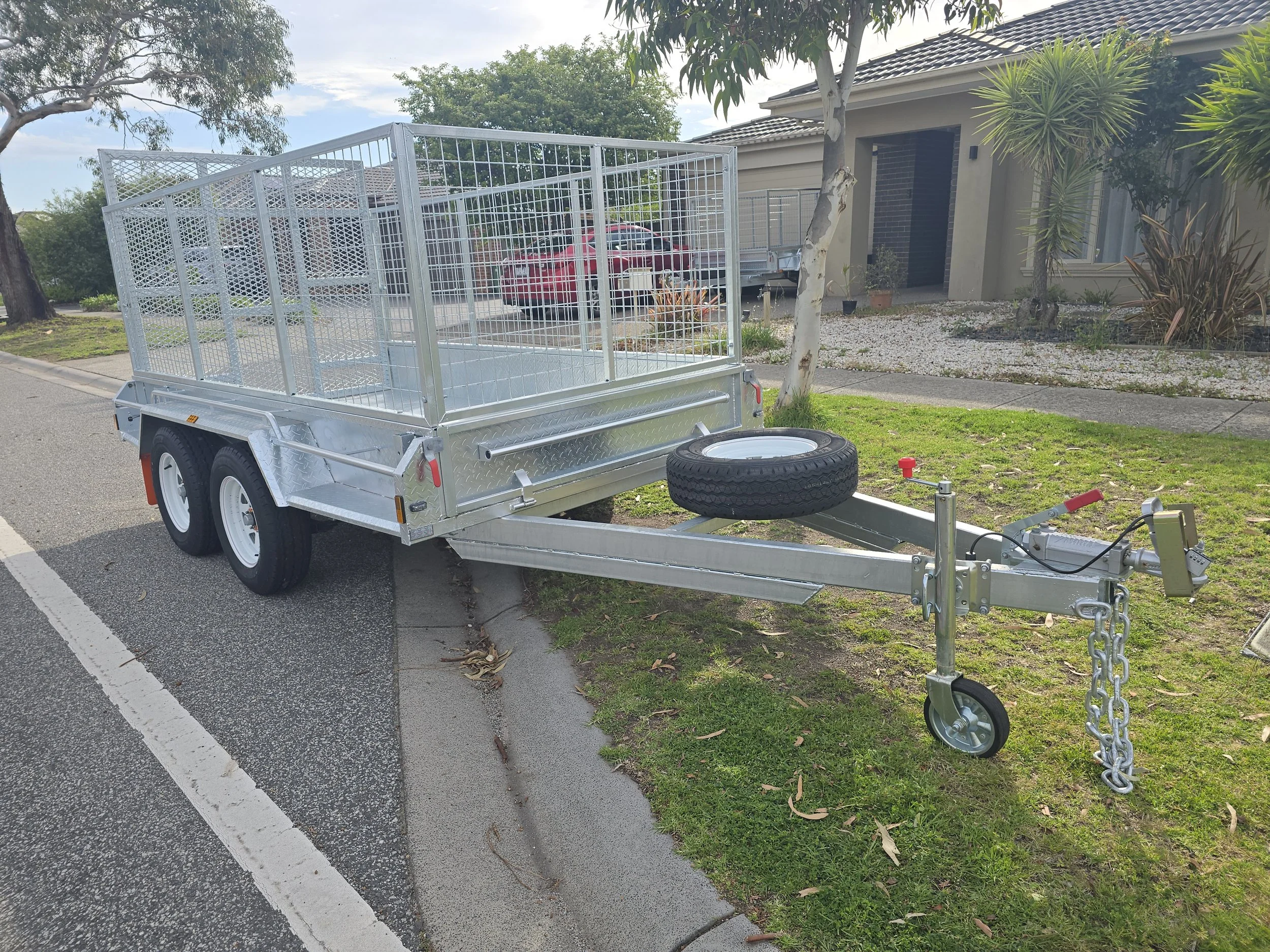 A silver metal utility trailer with a mesh cage, parked on a residential street next to a grassy area with trees, and a house with a tiled roof in the background.