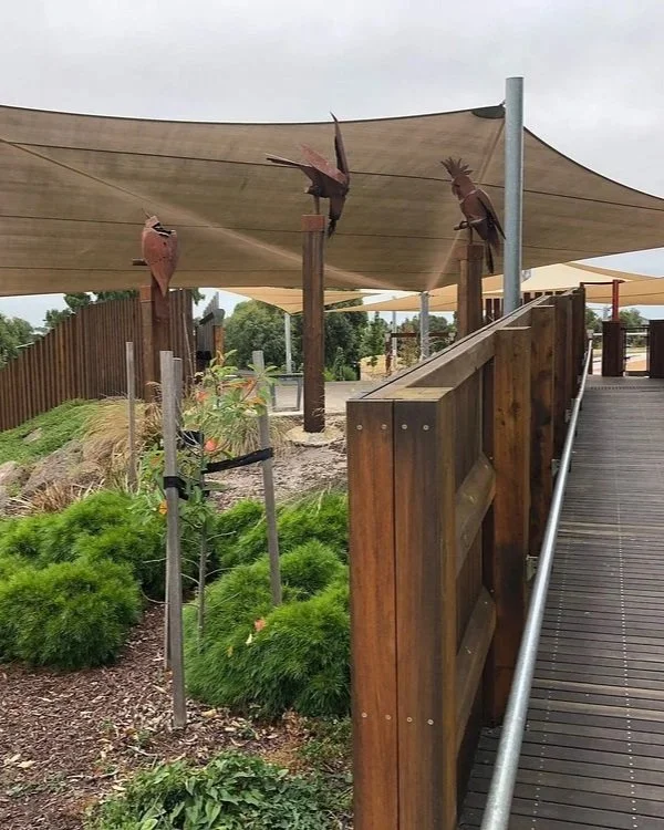 Three bird sculptures perched on wooden posts under a shade sail in a landscaped outdoor area with greenery and a wooden railing.