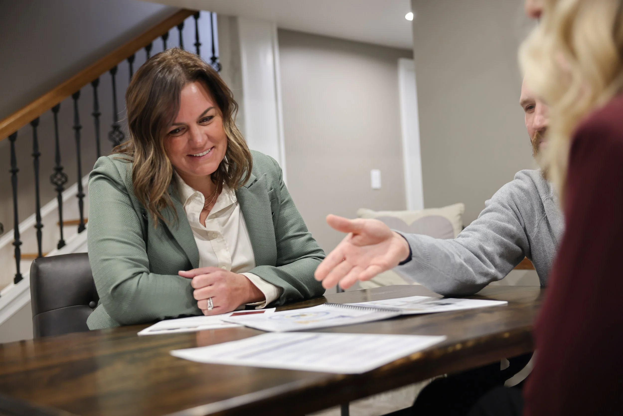 A woman in a gray blazer smiling during a meeting at a wooden table with papers, engaging with two other people.