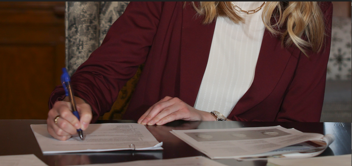A woman in a burgundy blazer and white blouse sitting at a desk, writing on a document with a blue pen, with additional papers and a newspaper on the desk.