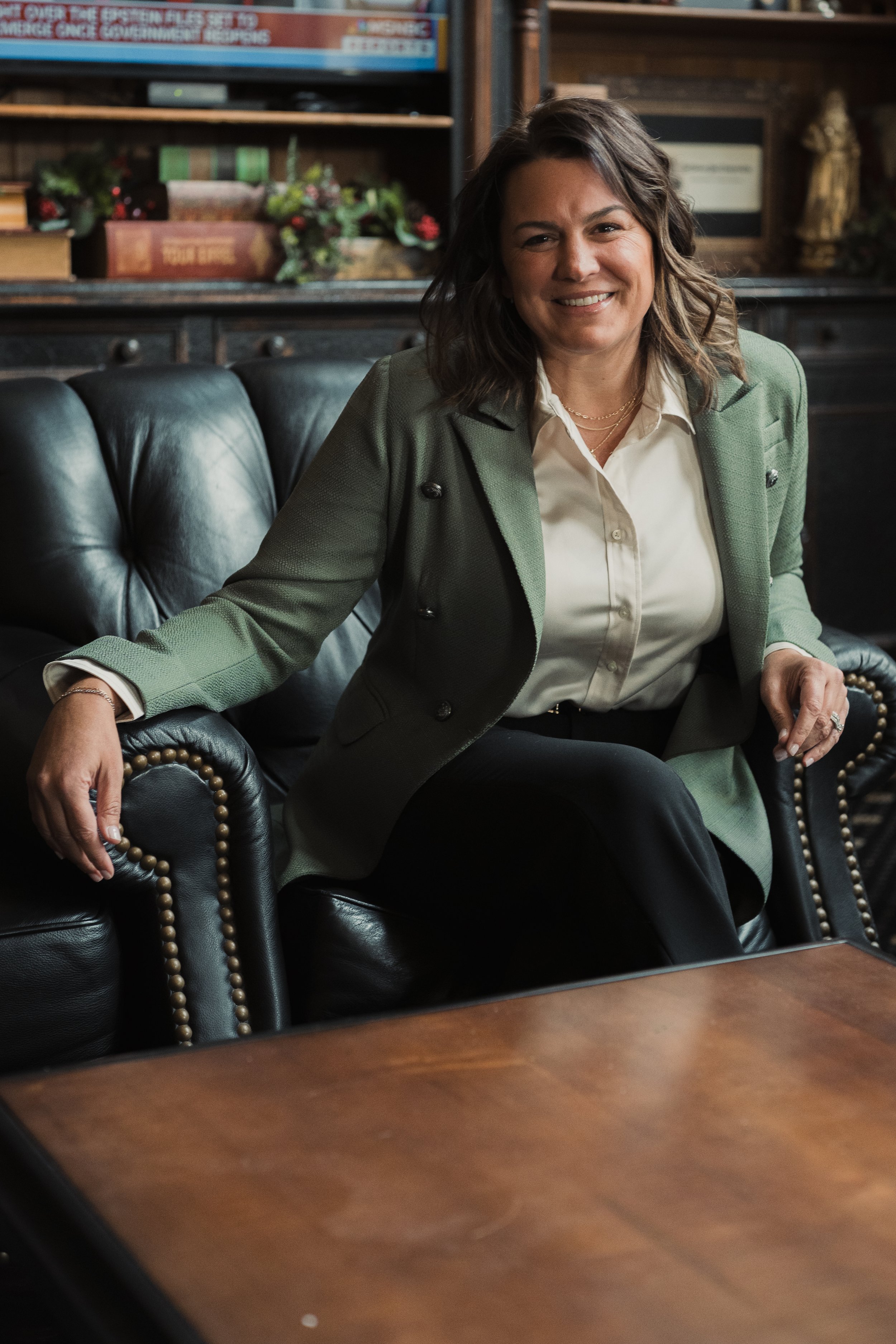 A woman with shoulder-length brown hair, smiling, seated on a black leather couch in a room with wooden bookshelves and decorative items. She is wearing a light green blazer and a cream blouse.
