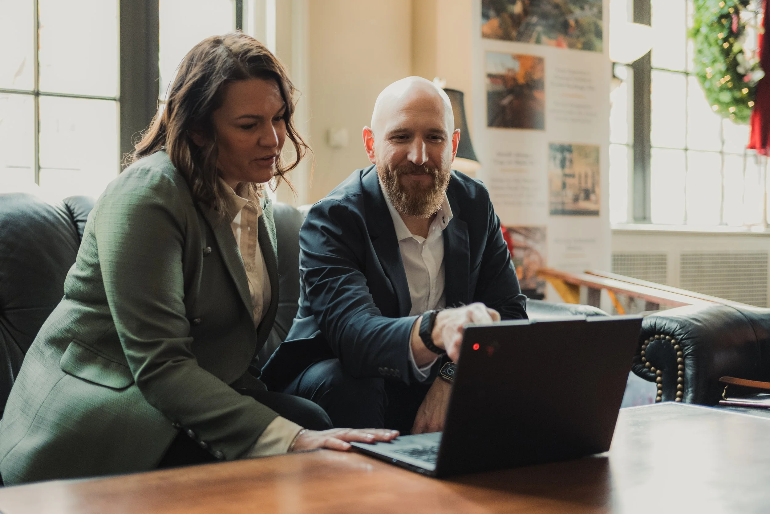 A man and a woman sitting on a couch, looking at a laptop on a table in a bright room with large windows.