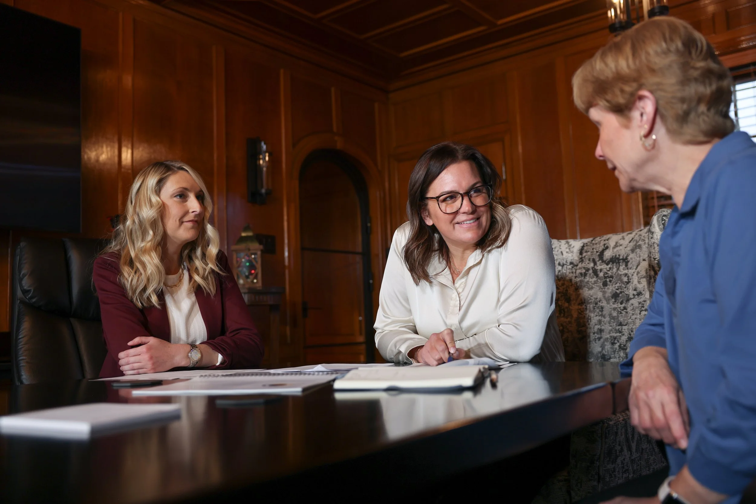 Three women having a conversation in a formal meeting room with wood-paneled walls.