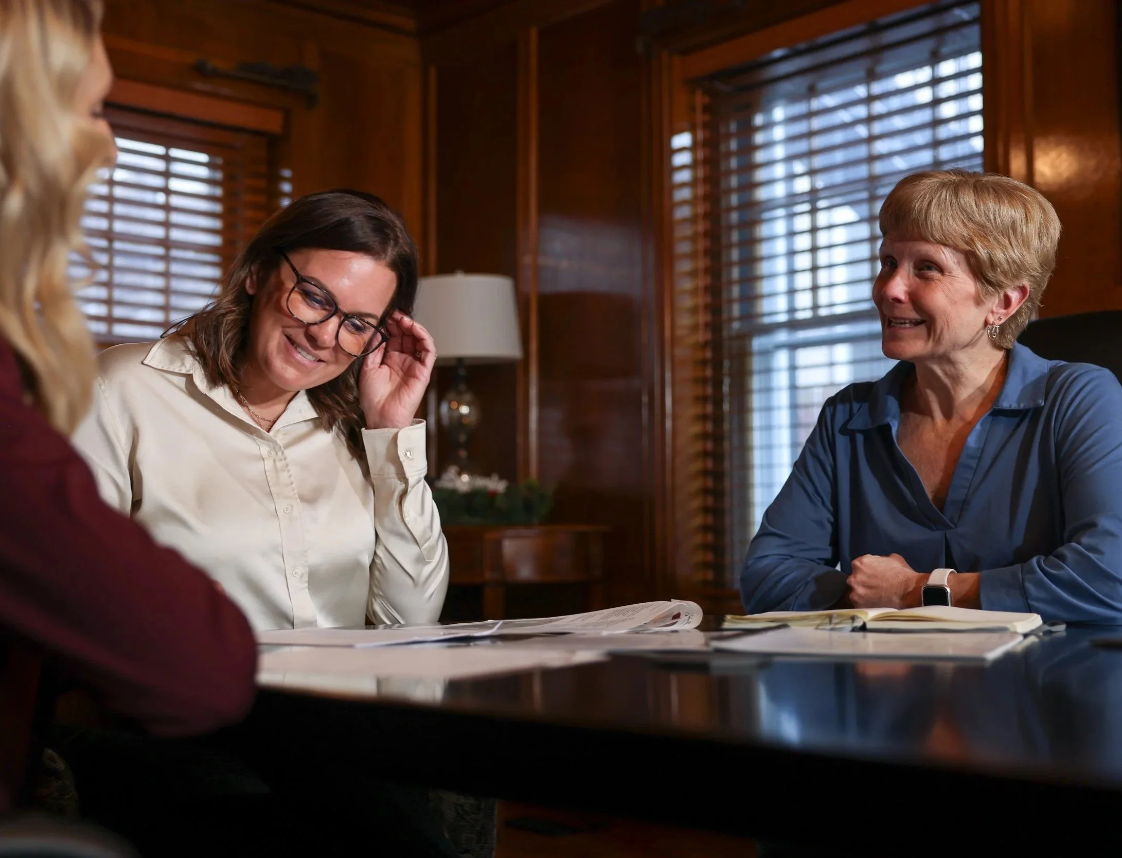 Three women sitting at a wooden table in a cozy wood-paneled room, engaged in conversation and smiling. The woman on the right has short blond hair and is wearing a blue shirt. The woman in the middle has glasses and brown hair, wearing a white shirt, and appears to be laughing or smiling.