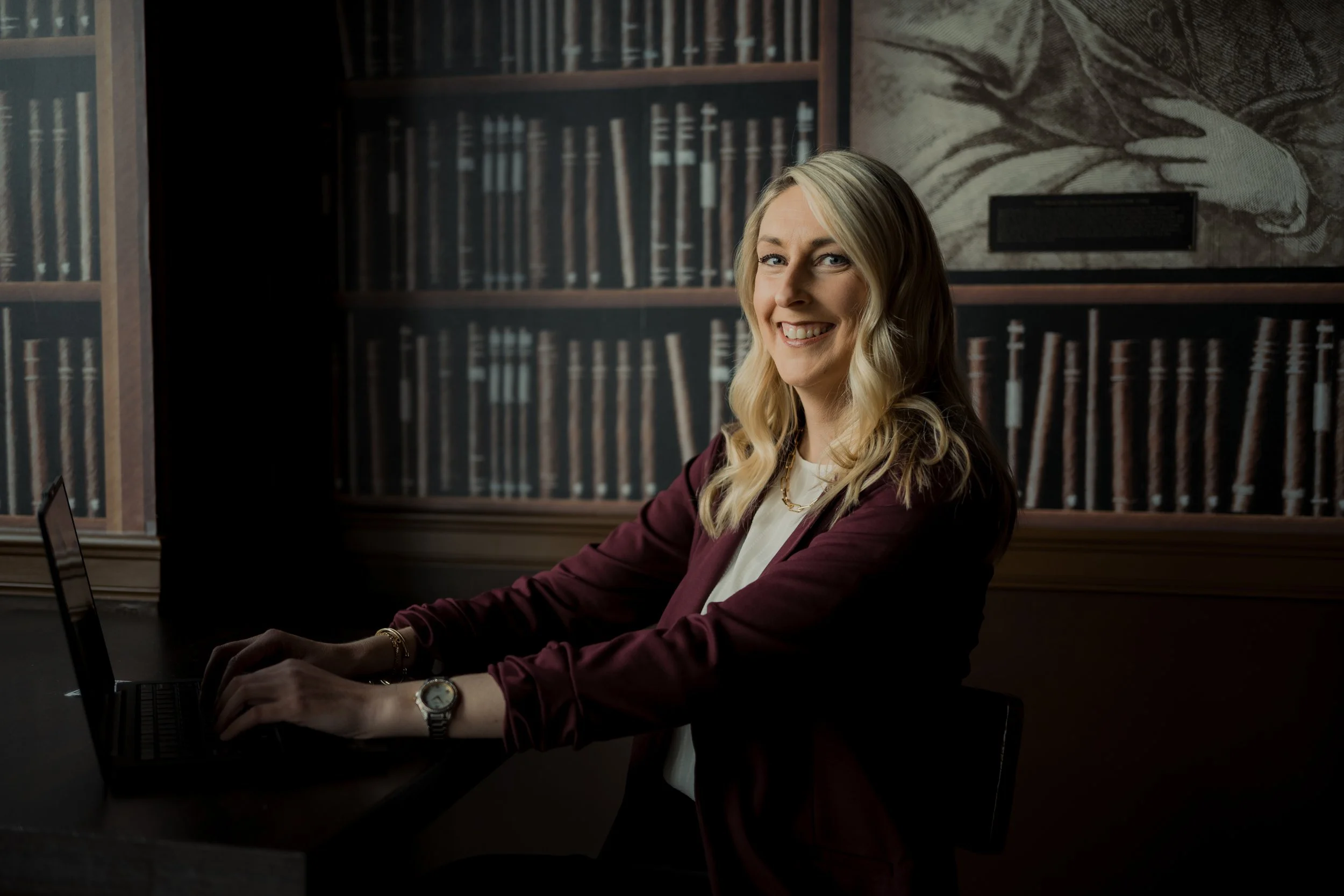 A smiling woman with blonde hair working on a laptop in a library setting.