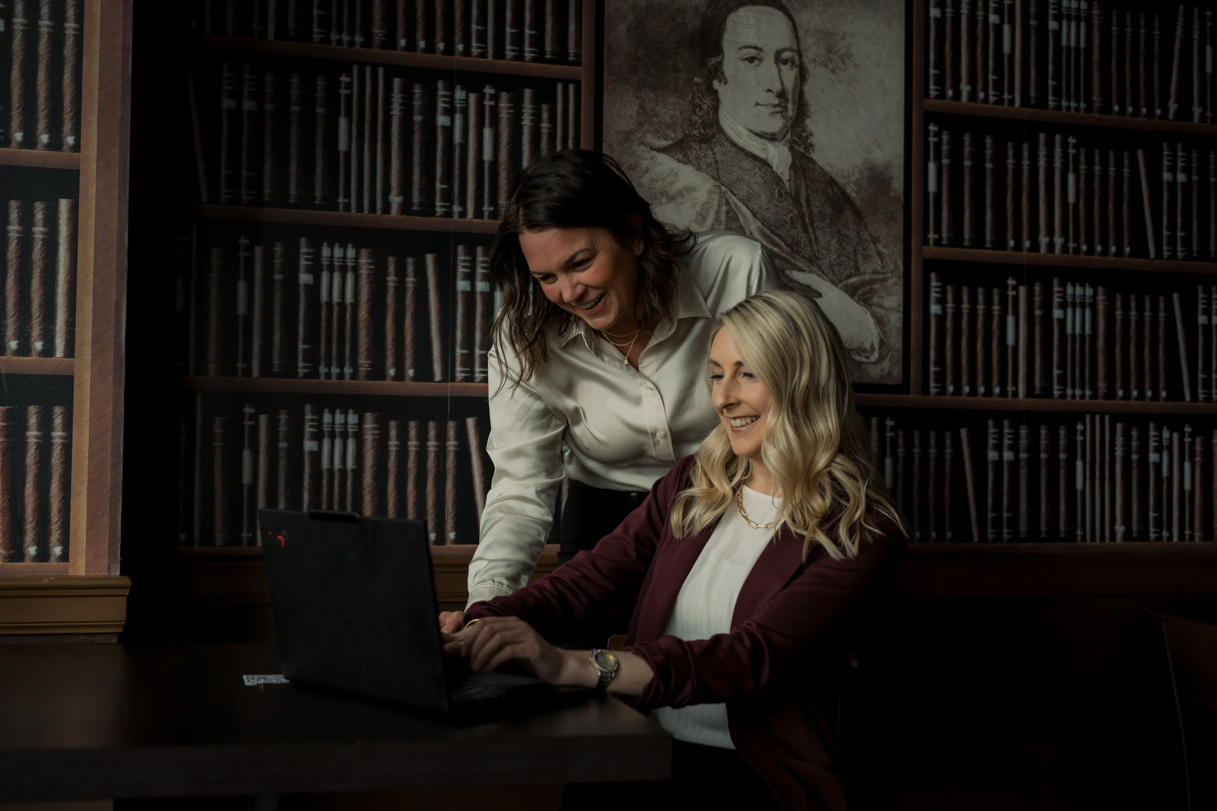 Two women working together on a laptop at a table in a library with wooden shelves and a historical painting in the background.