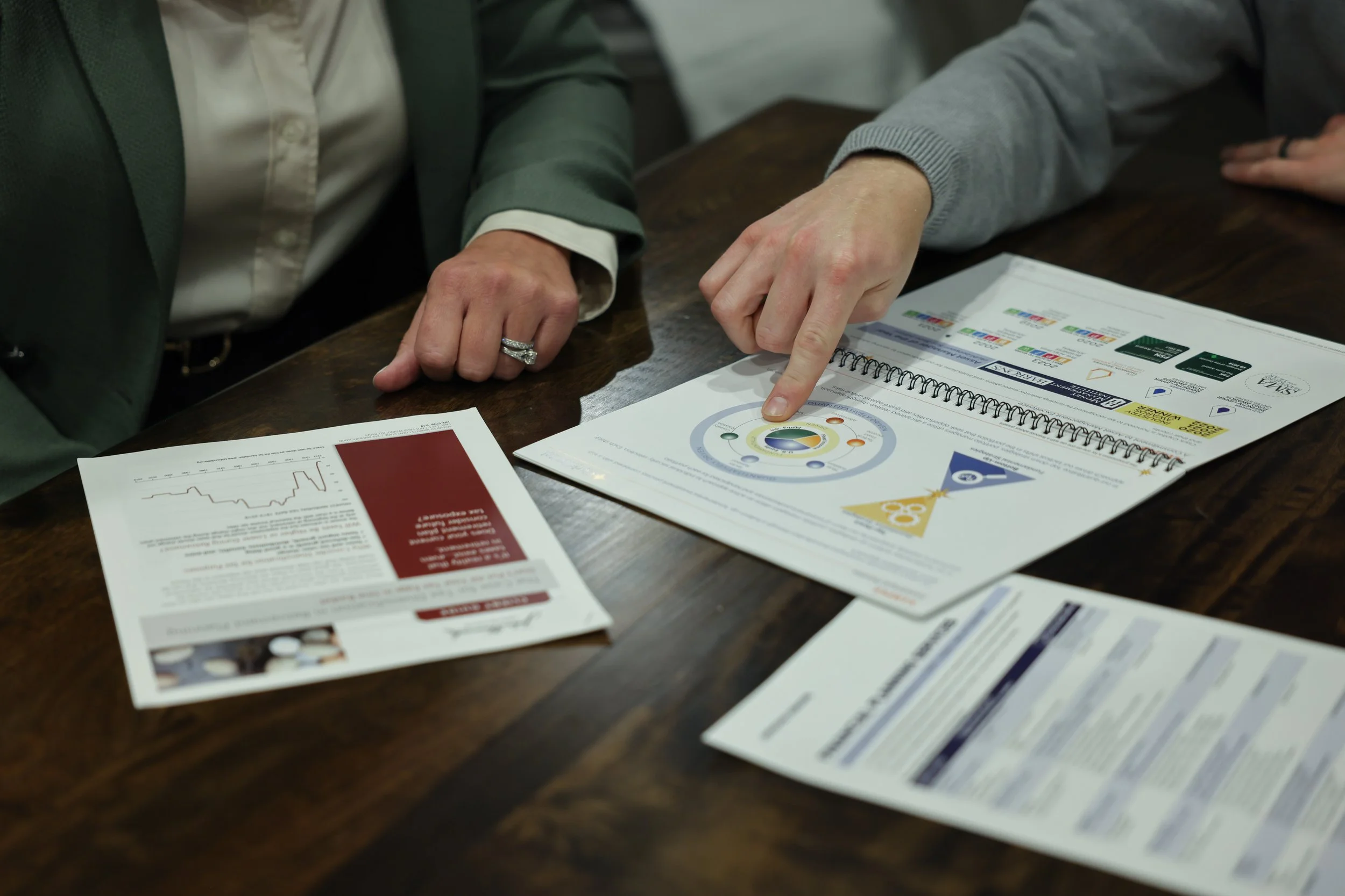 Two people discussing documents at a wooden table. One person is pointing at a colorful infographic in a spiral-bound booklet, while the other has a printed report with graphs and text.