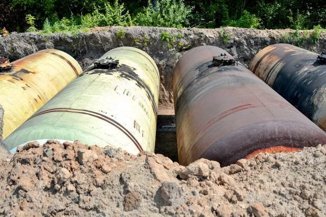 Four large, weathered underground storage tanks partially buried in a dirt excavation site, with greenery and trees in the background.