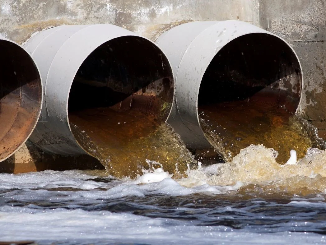 Three large metal pipes releasing brownish water into a body of water.