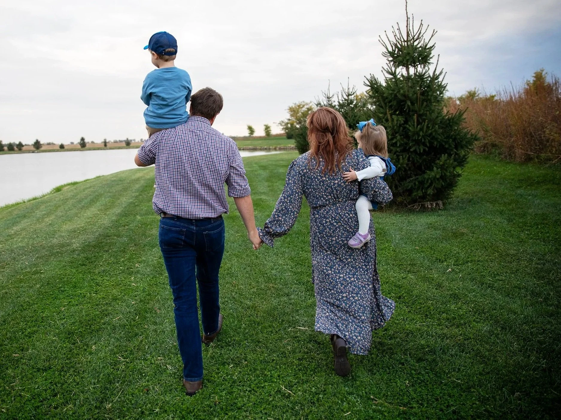A family of four walking outdoors on a grassy path next to a lake. The father is holding a young boy on his shoulders, the mother is holding the hand of a young girl, and they are walking towards a Christmas tree near bushes and trees.