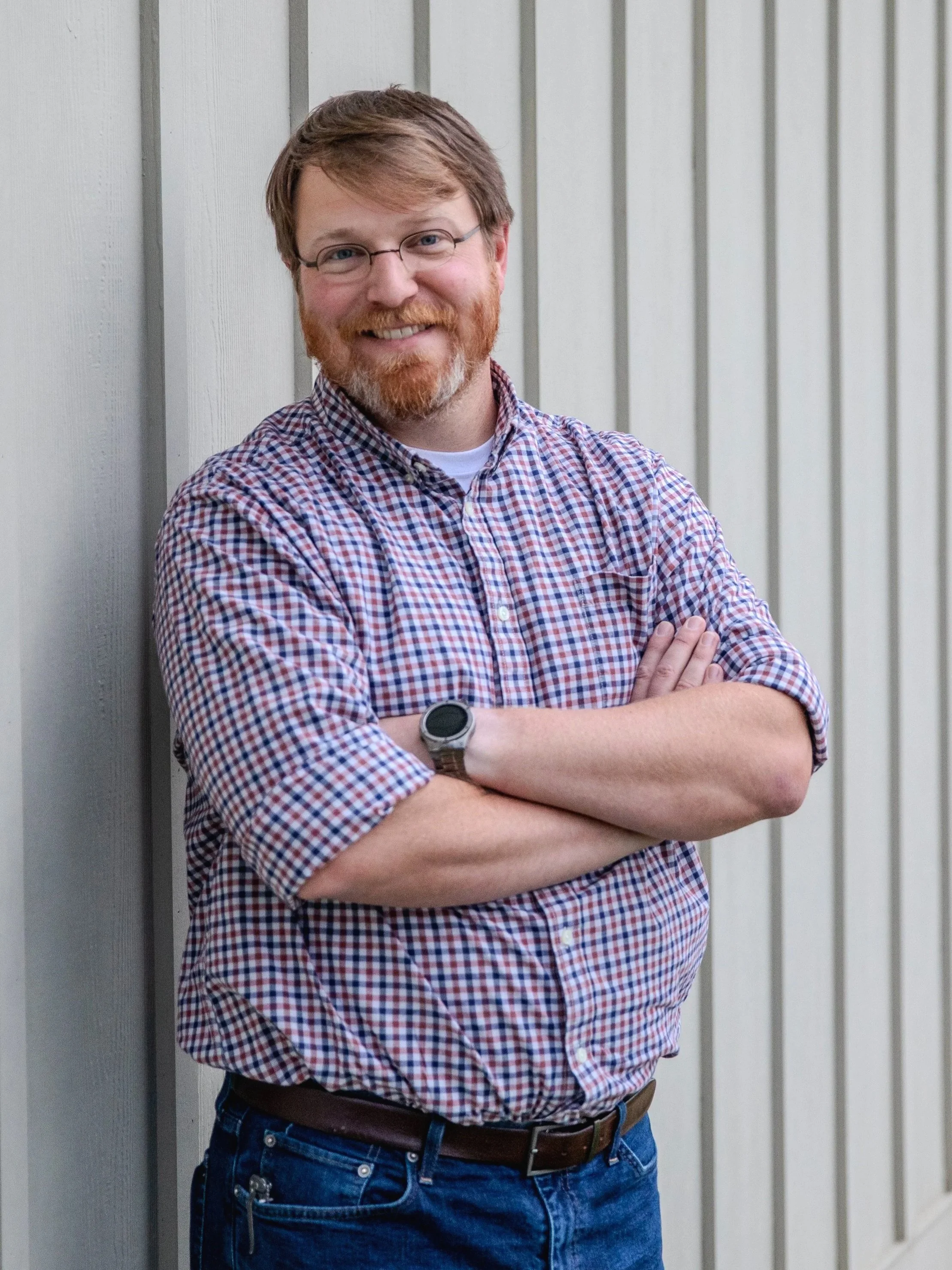 A man with glasses, a red beard, and mustache standing with arms crossed in front of a gray wooden wall.