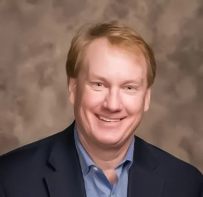 A smiling man with light skin and light brown hair, wearing a dark suit jacket and a blue collared shirt, against a neutral background.