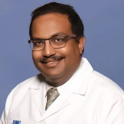 Portrait of a man wearing glasses, a white medical coat, and a tie, smiling against a blue background.