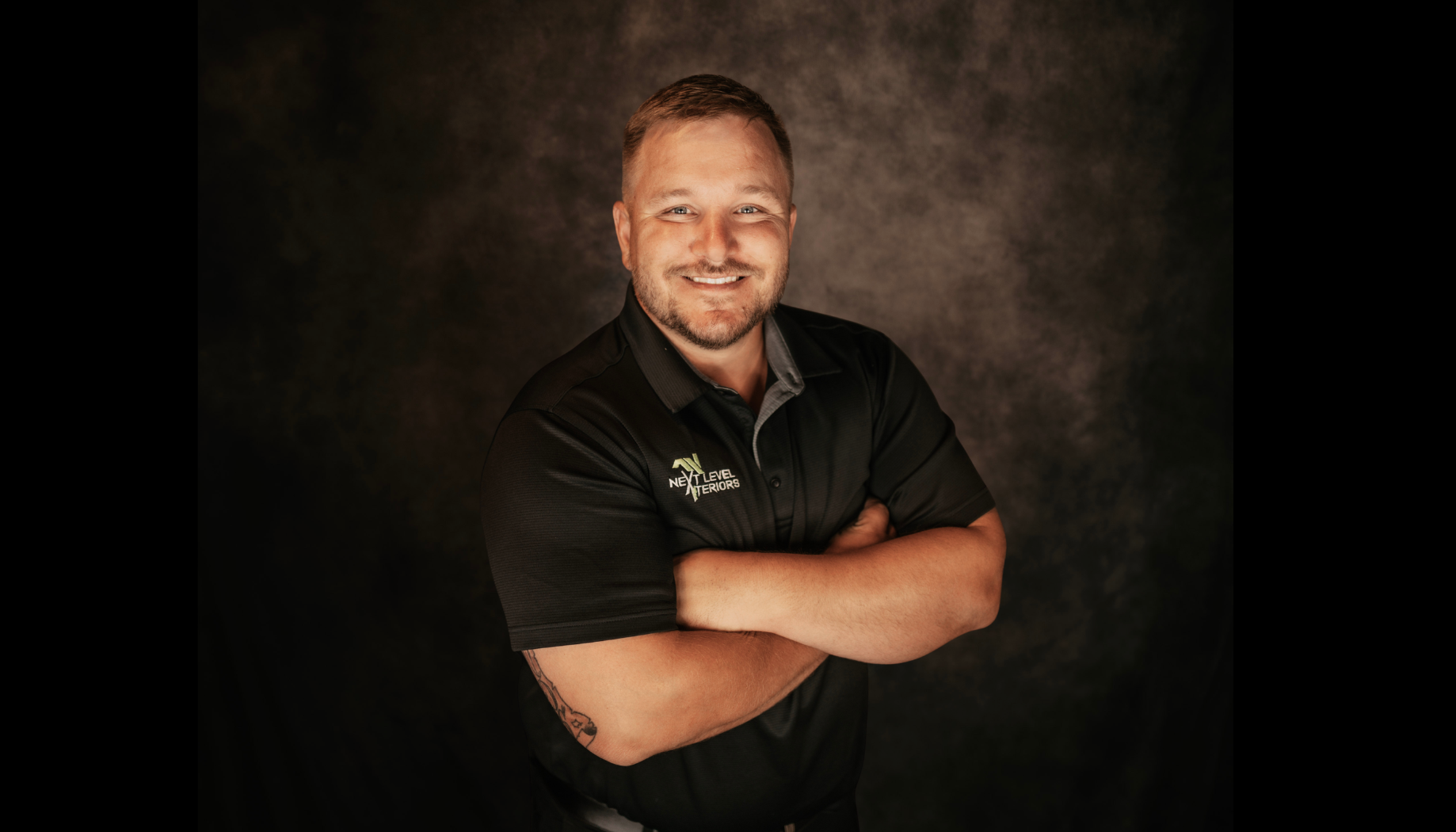 Portrait of a smiling man with arms crossed, wearing a black polo shirt with a logo, standing against a dark background.