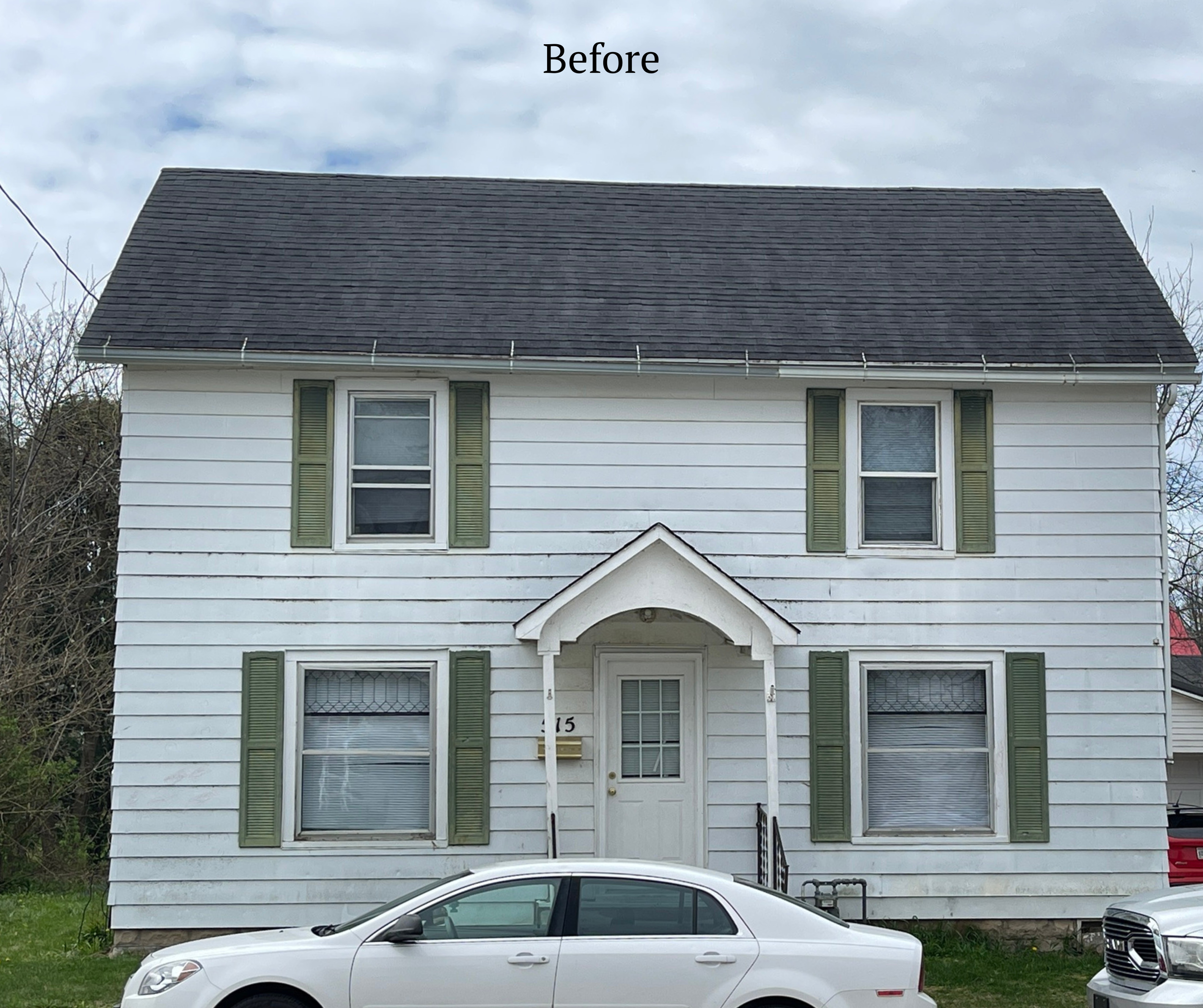 Front view of a two-story white house with green window shutters and a small arched porch over the front door, with parked cars in front and cloudy sky above.
