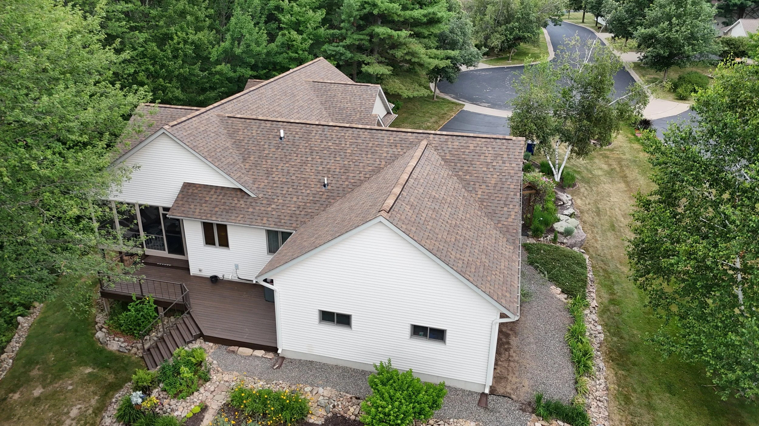 Aerial view of a white house with a brown shingle roof, surrounded by trees, a manicured lawn, and a curved driveway.
