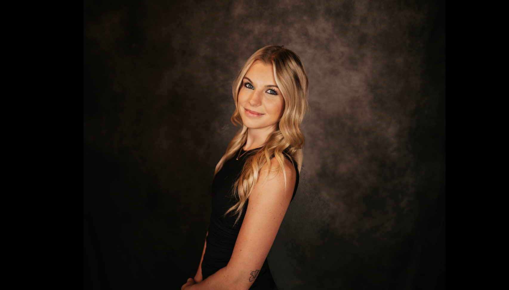 Young woman with long blonde wavy hair, blue eyes, wearing a black sleeveless top, smiling, posing against a dark, textured background.