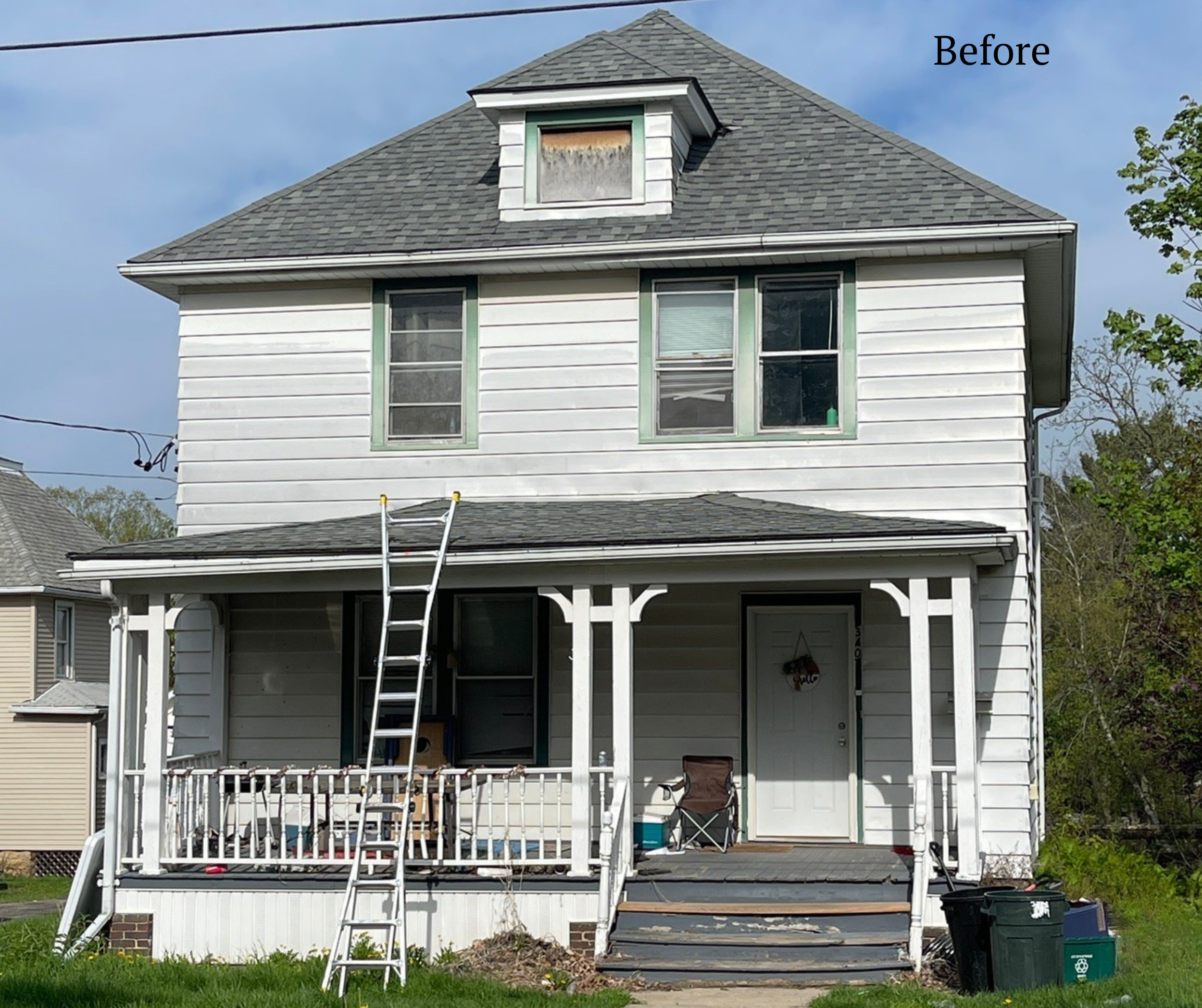 Before photo of a white two-story house with a front porch, including a ladder leaning against the house, and an old attic window covered with plywood.