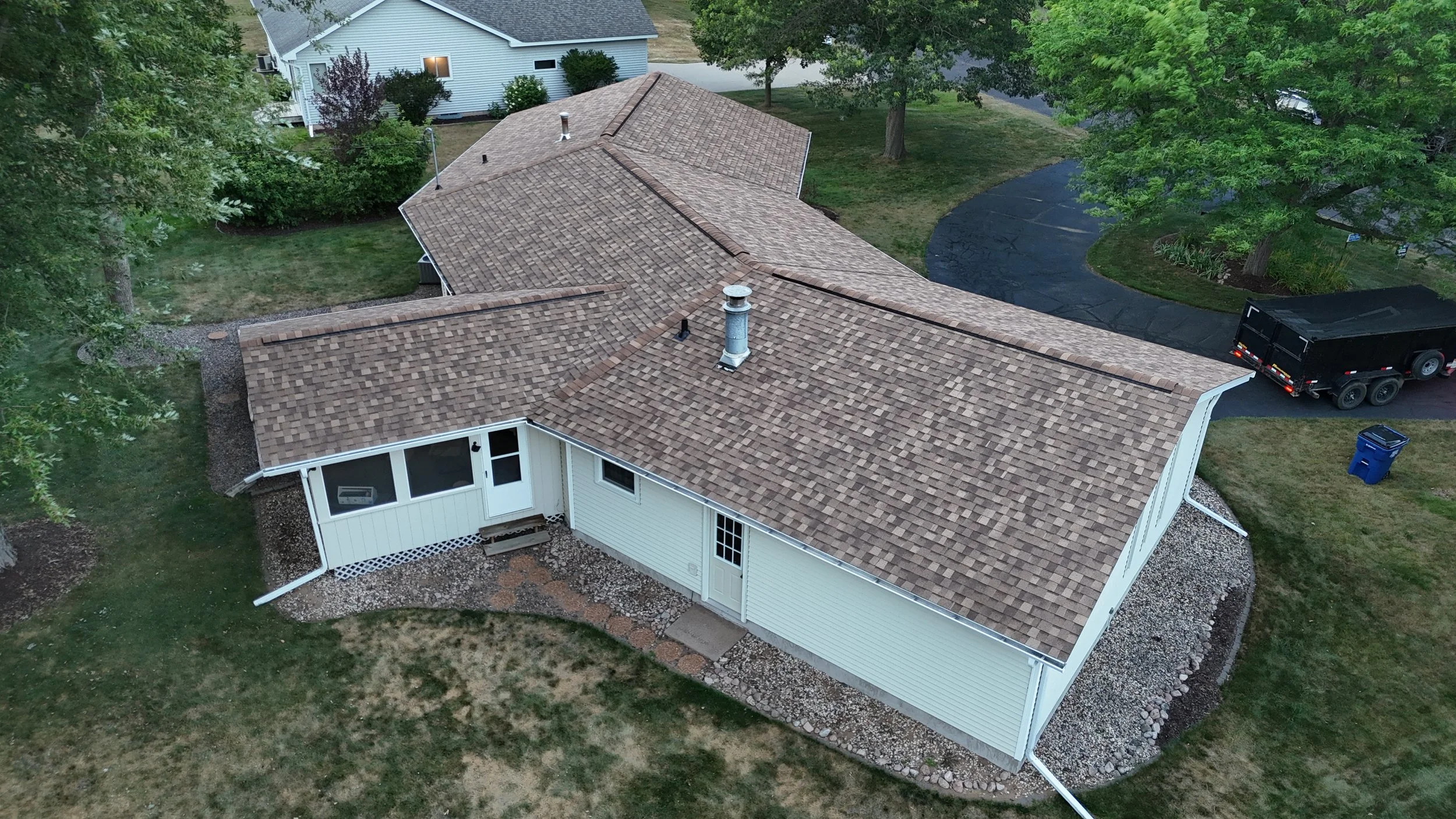Aerial view of a house with a tan shingled roof, surrounded by green trees and a lawn, with a black trailer parked on the driveway and a blue trash bin nearby.
