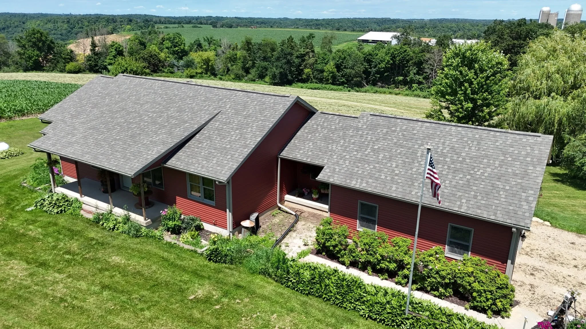 Red house with gray shingle roof, front porch, and American flag on a pole, surrounded by green lawn and garden with trees and fields in the background.